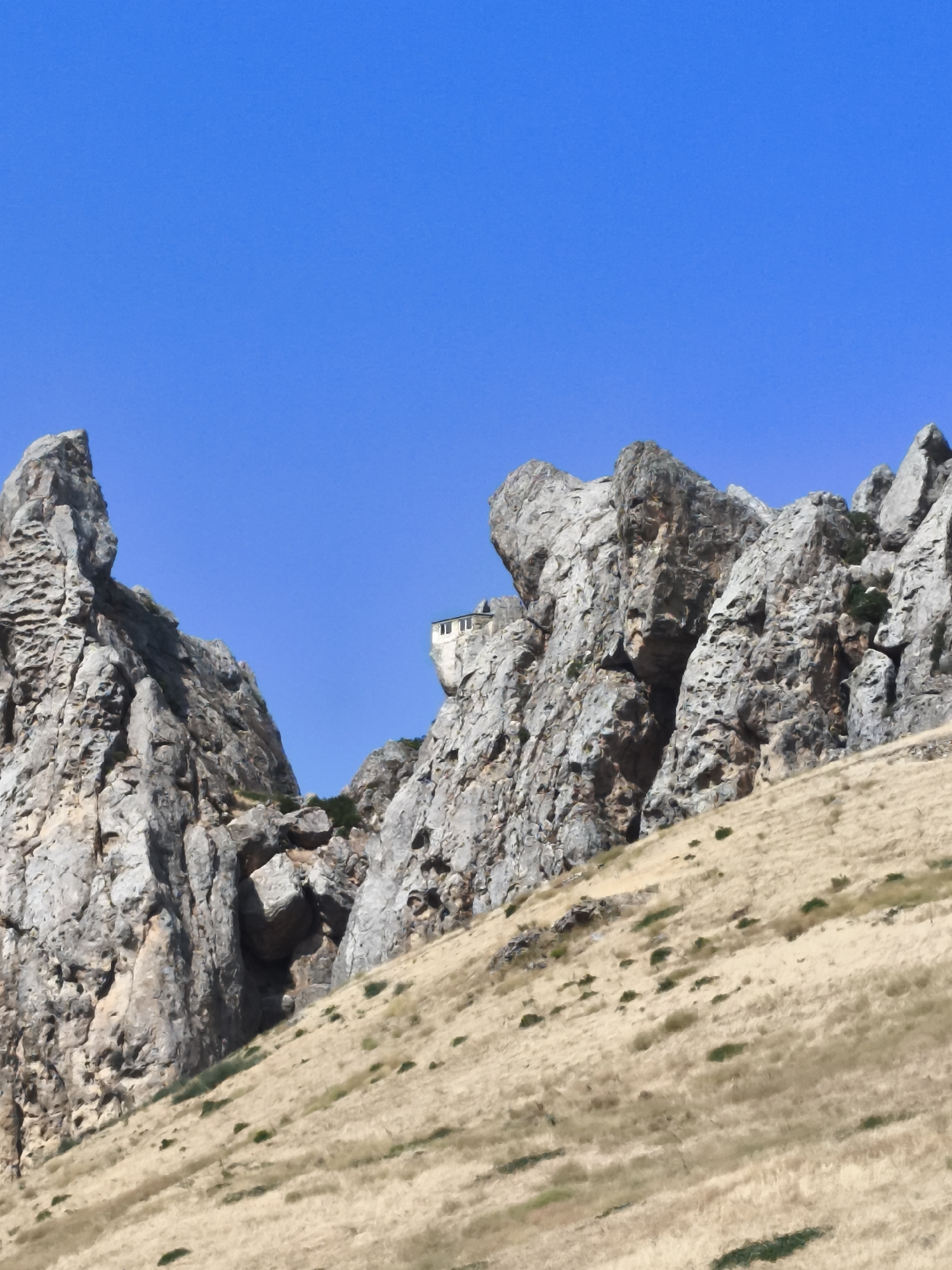 The image depicts a rugged, mountainous landscape under a clear blue sky. In the foreground, a dry, grassy slope rises towards imposing rock formations. These rock formations are characterized by their rough, weathered surfaces and dramatic contours, with one prominent peak on the left and a cluster of jagged rocks on the right. Perched precariously on a ledge of the rock formation on the right is a small, white building with a dark roof, appearing to be a monastery or a hermitage. The overall environment suggests a remote and arid location, typical of mountainous regions in Azerbaijan, potentially near the city of Siazan known for its geological formations. There are no people visible in the image.