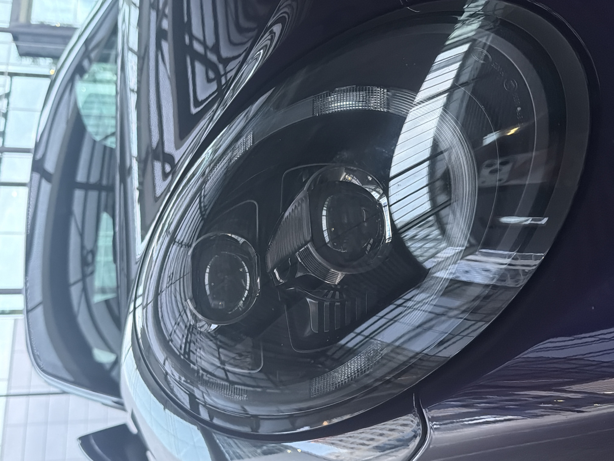 A close-up, low-angle shot captures the sleek, dark blue exterior of a Porsche 911, focusing on its distinctive round headlight assembly. The car is parked indoors, likely in a showroom or garage, as evidenced by the reflective ceiling with a grid pattern of lights and beams. The glossy finish of the car's bodywork mirrors the surrounding architecture, creating a dynamic interplay of light and shadow. The headlight itself is a complex piece of engineering, featuring multiple circular projectors and intricate detailing within its clear lens. The image is sharp and detailed, highlighting the car's sophisticated design and the quality of its construction.