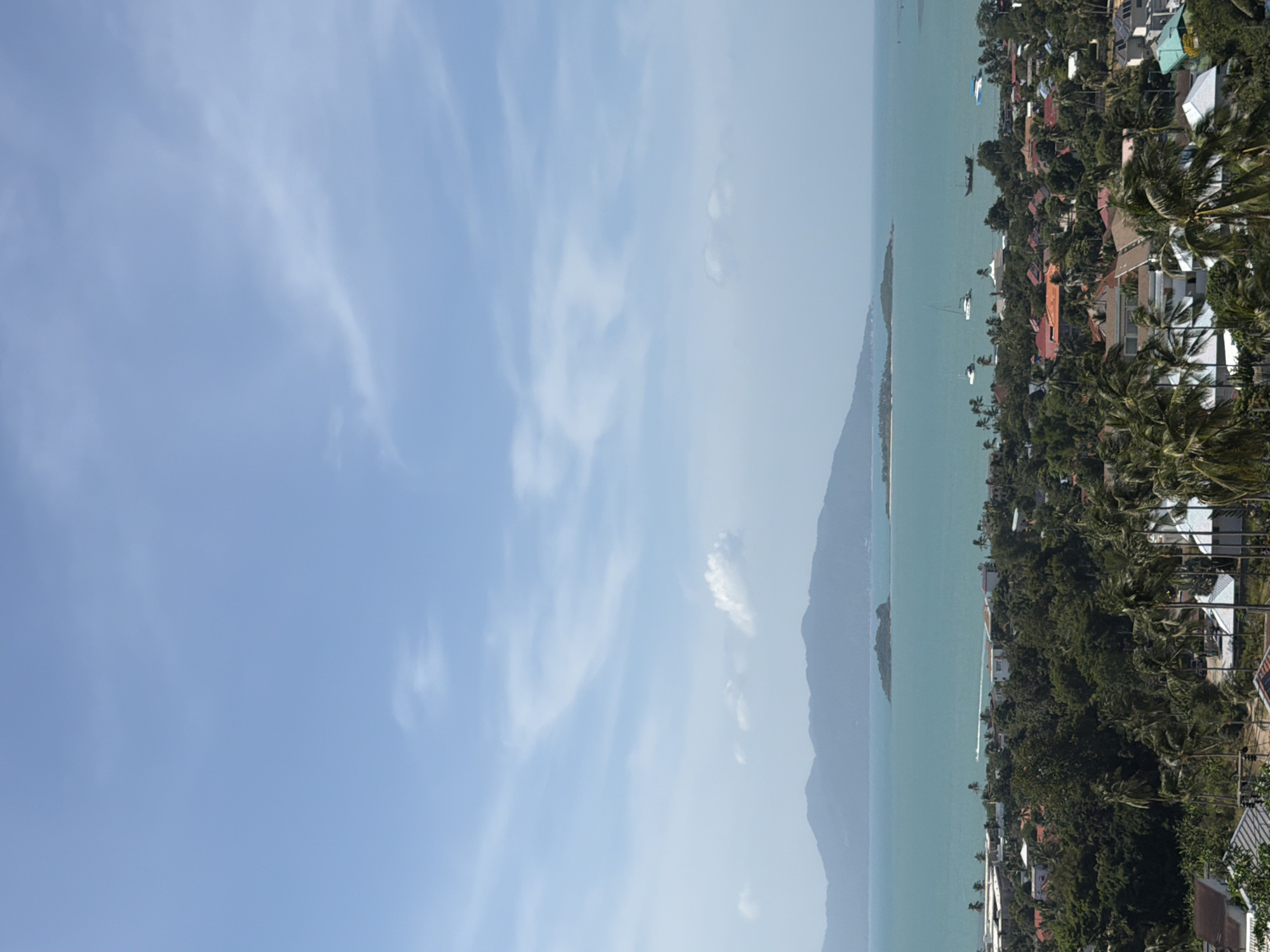 This image depicts a panoramic view of Ko Samui District, Thailand, on a clear, sunny day. The foreground is dominated by lush green foliage, including numerous palm trees, interspersed with residential buildings with diverse roof colors, ranging from red and orange to white and gray. A few of these buildings feature white walls and white tiled roofs.
In the mid-ground, a dense canopy of trees forms a verdant landscape that stretches towards the coast. Beyond the trees, a town or village is visible with a collection of buildings, some with orange-tiled roofs and white walls. Several boats, including sailboats, are dotted across the turquoise water.
The background features the expansive turquoise sea, beyond which lie several islands and a large, mountainous landmass in the distance, its peaks softened by atmospheric haze. The sky above is a vibrant blue, with scattered, wispy white clouds. A white boat with a wake trails across the water in the mid-distance. The overall impression is one of a tropical coastal community.