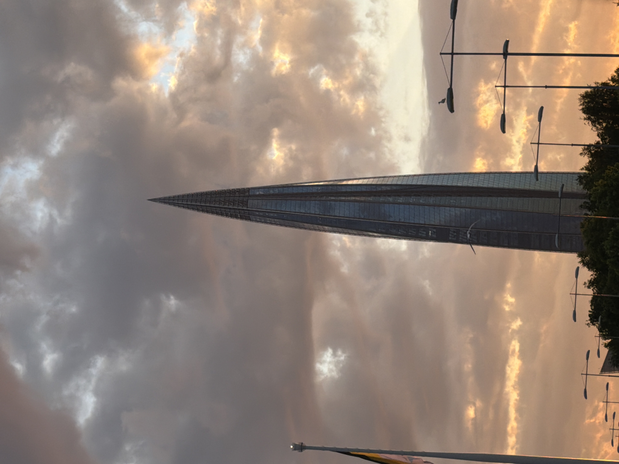 The image depicts the Lakhta Center, a towering, modern skyscraper, set against a dramatic sky in Saint Petersburg, Russia. The building, clad in reflective glass, tapers significantly as it rises, culminating in a sharp point. Its vertical lines and slightly spiraling form are clearly visible, dominating the left side of the frame.
The sky is a prominent feature, filled with dynamic clouds that show the characteristics of either sunrise or sunset. Patches of warm golden, orange, and yellow light pierce through darker grey clouds, indicating the sun is low on the horizon and partially obscured. This creates a striking contrast and an atmospheric glow across the scene.
On the right side of the image, several tall streetlights or utility poles with multiple extended arms are silhouetted against the bright sky. A small bird, possibly a seagull, is perched on one of these streetlight arms. Further to the right, the dark green foliage of trees is visible. Towards the bottom, a flagpole with a multi-striped flag (appearing dark and light, possibly black, yellow, and red) is partially visible. No people or specific activities are depicted. There is no visible text. The overall scene emphasizes the grandeur of the architecture against a powerful, naturally lit sky.