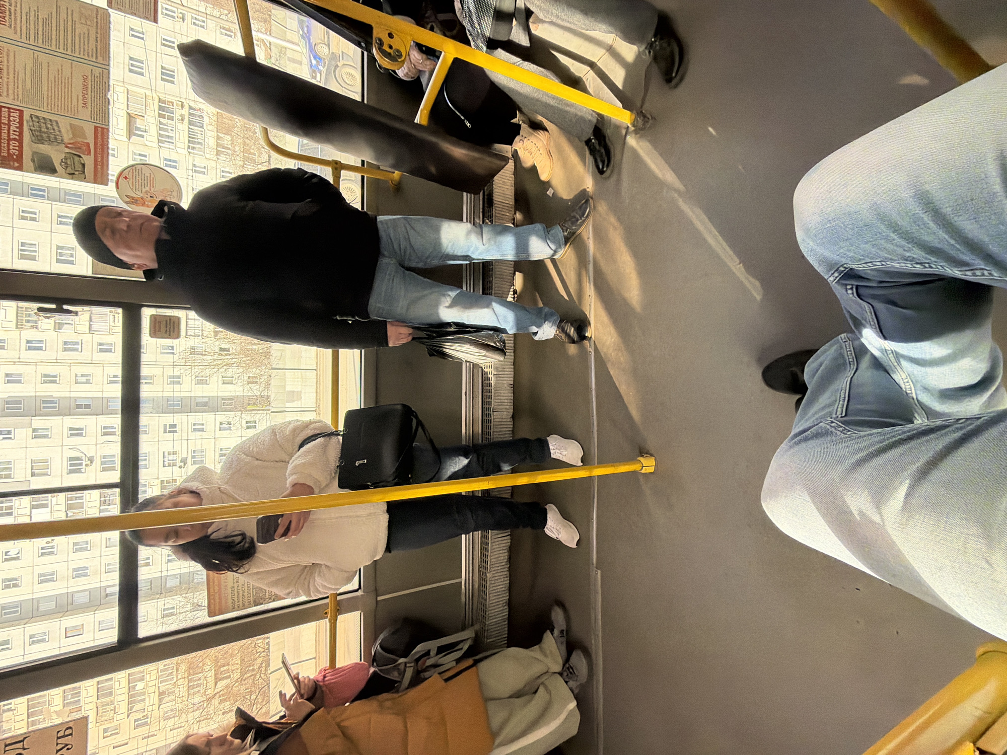 This media file captures the interior of a public transport vehicle, likely a bus or trolleybus, in Krasnoyarsk, Russia, during what appears to be a sunny day. The scene shows several passengers standing and sitting, illuminated by bright sunlight streaming through large windows. The image is taken from a slightly tilted, low-angle perspective.
In the foreground, a person's legs clad in light blue jeans are visible, suggesting the viewpoint of a seated passenger. To the left, a man in a black jacket, light blue jeans, and a black beanie stands, holding a black umbrella. He looks forward, his face partially visible. Behind him, another passenger, a woman in a white fluffy jacket and dark pants, holds a smartphone. Her face is also partially visible, looking down. Both are holding onto yellow grab poles, a common feature in public transport. Further down, another person with an orange jacket and a pink hat is seated, engrossed in their phone. To the right, other passengers' legs are visible, some seated, some standing, wearing various shoes including black and beige.
Through the windows, typical multi-story residential buildings, characteristic of Russian urban architecture, are visible. The strong sunlight casts sharp shadows inside the bus.
Several pieces of Russian text are visible on signs affixed to the windows and interior walls. These include:
* "ВОДИТЕЛЕЙ на маршруты №12" (Drivers for routes #12)
* "МОТОРИСТА" (Mechanic)
* "АВТОЭЛЕКТР" (Auto Electrician) – indicating job advertisements.
* "БЕСКОЗНЫЕ ВЕЩИ - ЭТО УГРОЗА!" (Unattended items - this is a threat!) – a safety warning.
* "ЗАПРЕЩЕНО" (Forbidden/Prohibited) – likely related to rules or safety.
* "ПАМЯТ" (likely part of "Памятка" meaning memo or instruction).
The overall impression is one of routine daily commute in a Russian city.