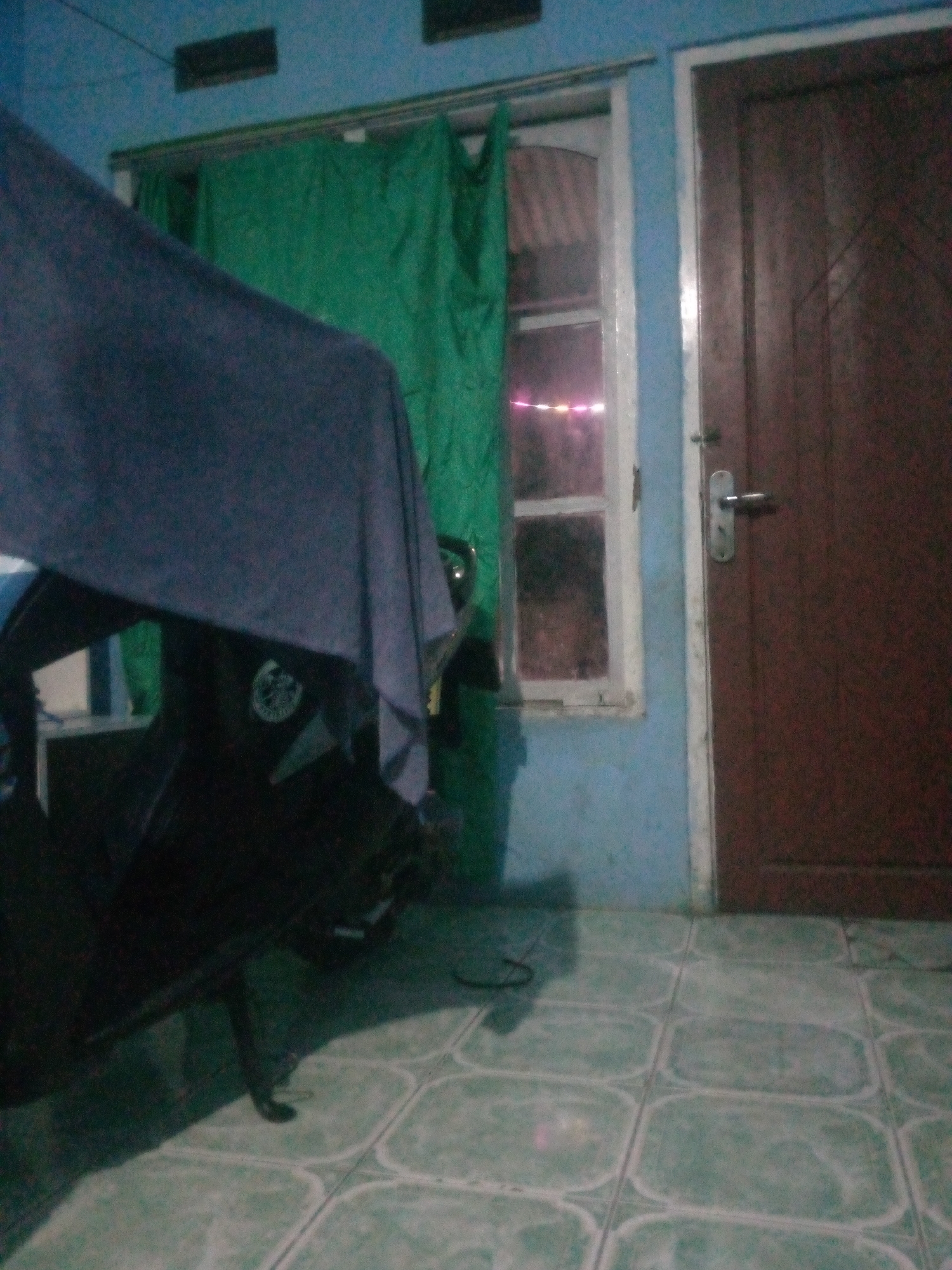 The image depicts an indoor scene, likely a garage or covered area, with a motorcycle partially covered by a dark blue cloth in the foreground. To the left of the motorcycle, a portion of a wall painted a light blue is visible. In the background, a green curtain hangs in front of a window. To the right, a brown wooden door with a silver handle is present. The floor is covered with light green tiles. The overall lighting suggests it is nighttime or a dimly lit interior. The scene appears to be located in Giriasih, Indonesia, as stated in the prompt.