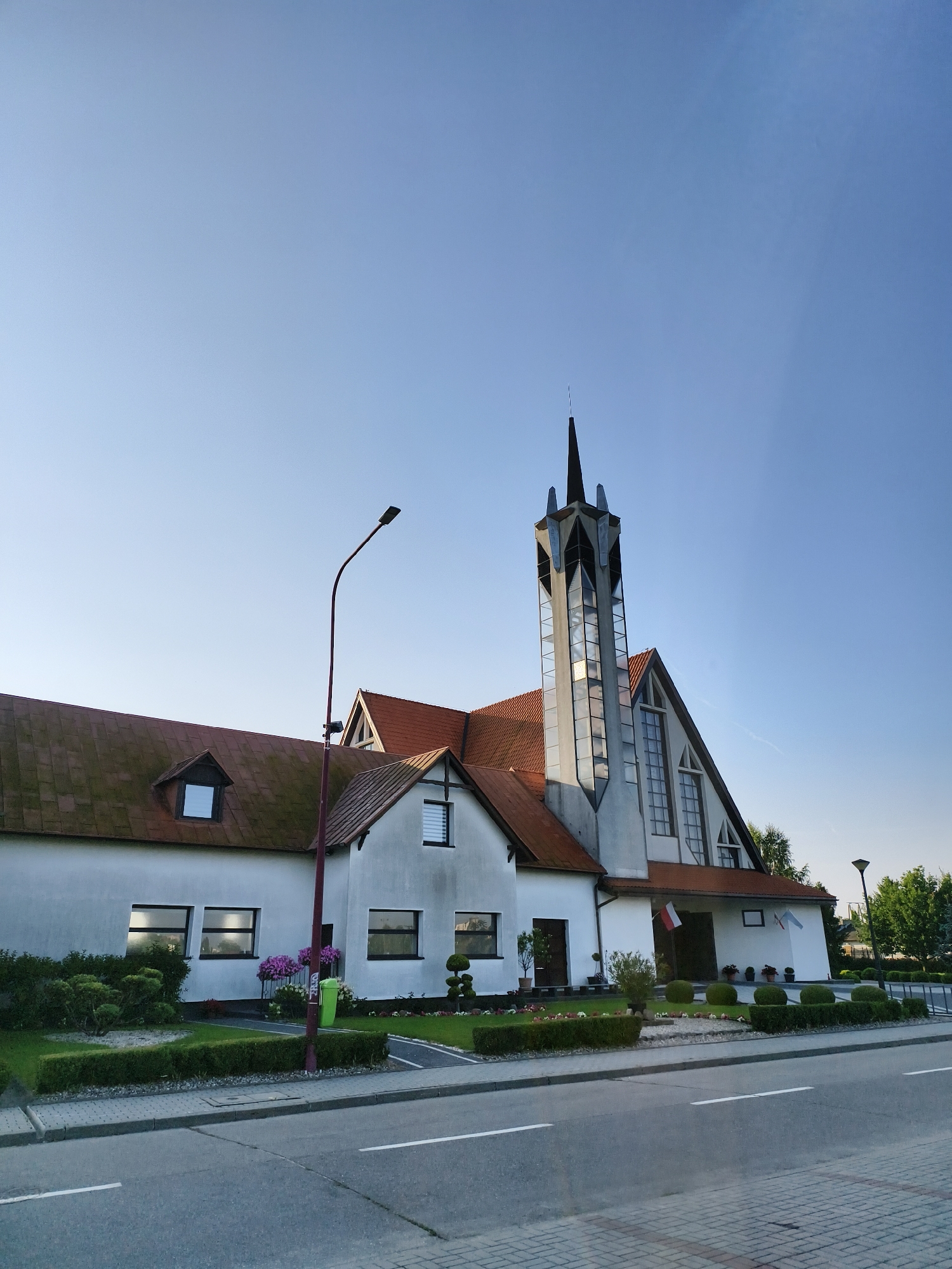 The image shows a modern church building with a distinctive tall, glass-paneled tower topped with a pointed spire. The church has a white facade with a red-tiled roof. Flags, one of which appears to be the Polish flag, are displayed near the entrance. The building is situated beside a road, with a sidewalk and manicured landscaping, including hedges and small trees, in front. A street lamp is visible to the left of the tower. The sky is clear and blue. The scene appears to be a daytime view in Tczew, Poland, capturing the exterior of the religious institution.