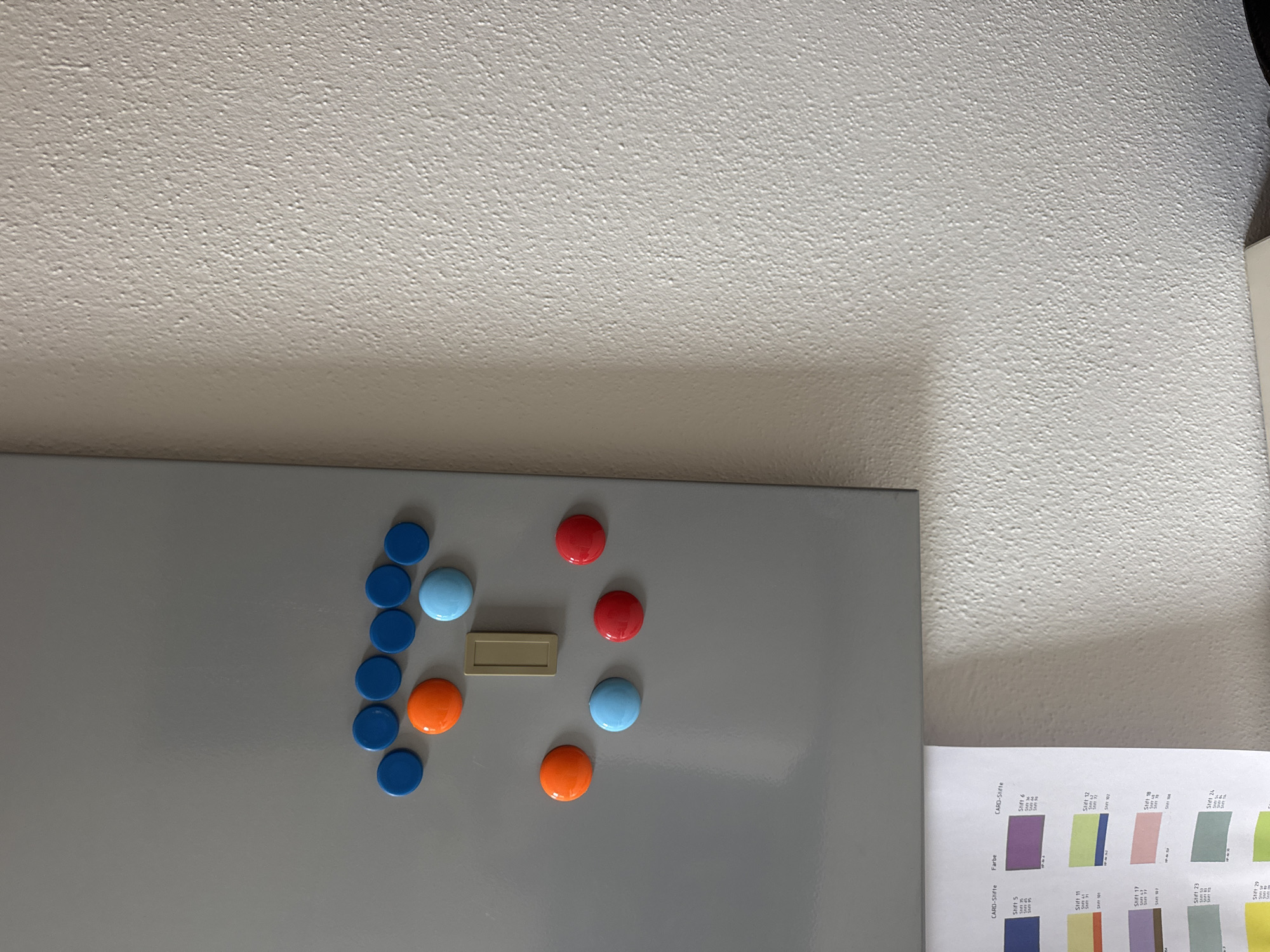 The image displays a gray magnetic board with several round magnets attached. The magnets are of various colors: blue, orange, red, and light blue. One rectangular beige object is also attached to the board. In the lower left corner, a portion of a document with color swatches and text is visible. The magnetic board is mounted on a textured, light-colored wall. The scene appears to be an indoor setting. There is no indication of the specific location within Hohenbrunn, Germany.