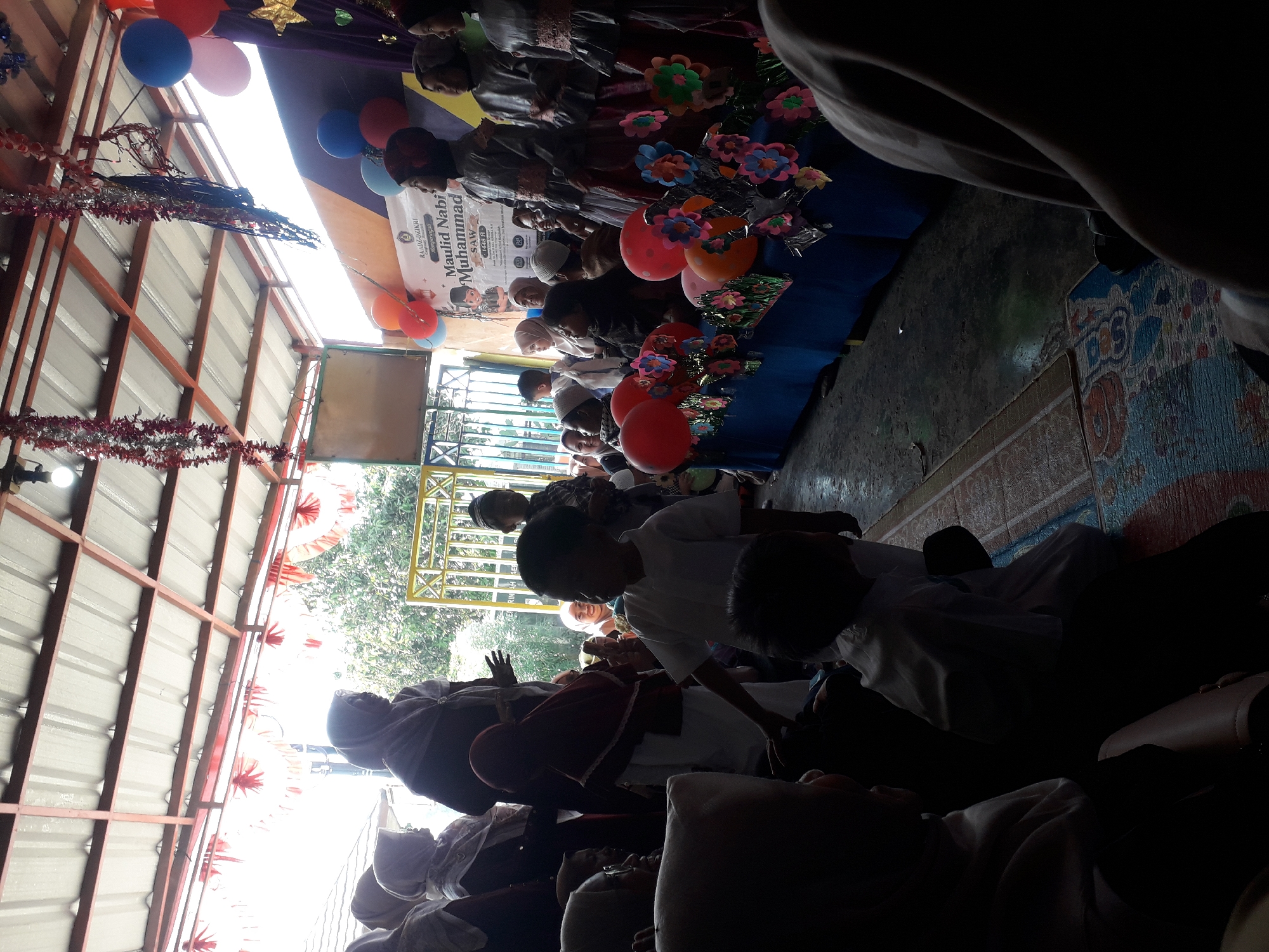 The image depicts an outdoor gathering, possibly a cultural or religious event, in Cankuang, Indonesia. A group of people, predominantly children and women wearing hijabs, are assembled under a roofed structure with a corrugated metal ceiling. In the background, a yellow metal gate and lush greenery suggest an outdoor setting. A banner with Indonesian text, including "Maulid Nabi Muhammad SAW," indicates the event commemorates the birthday of the Prophet Muhammad. Colorful balloons and paper flowers adorn a decorated table in the foreground, suggesting a festive atmosphere. Some individuals are interacting, while others are observing. The lighting suggests it is daytime, with sunlight filtering through the roof. The ground is covered with mats and a dark, textured surface.
