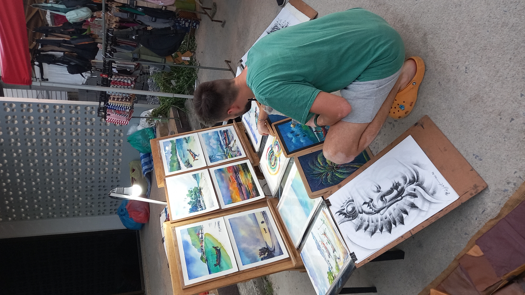 In Ko Pha-ngan District, Thailand, a man in a green t-shirt and grey shorts is crouched on a concrete surface, examining artwork. He is wearing yellow Crocs. In front of him is a display of paintings and drawings, primarily watercolors of tropical scenes and a graphite portrait of a face with elaborate headwear. Behind him, a rack of clothing and bags is set up under a red awning. Several colorful bags and a lit lamp are also visible in the background, suggesting this is an outdoor market or street stall.