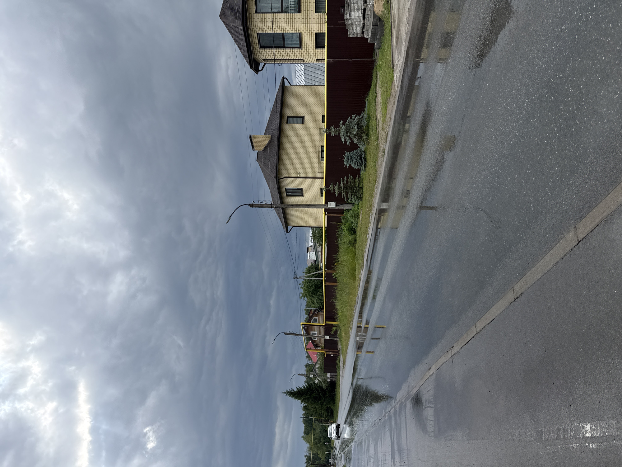 The image depicts a residential street in Kazan, Russia, on a cloudy day, likely after recent rainfall. The scene is dominated by a wet asphalt road reflecting the overcast sky and the surrounding buildings. On the left side of the road, there are two modern houses constructed with light yellow brick and dark brown roofs. They are separated by a dark brown fence. Small, conical evergreen trees are planted in front of the fence. A street lamp stands near the foreground house.
Further down the street, more houses are visible, with red roofs and wooden facades. A yellow gas pipe runs along the street and up the side of one of the houses. The sky is filled with heavy, grey clouds, suggesting gloomy weather. The wet road, with large puddles, indicates that it has been raining recently or is still raining lightly. In the distance, a white car is seen driving on the wet road, its tires creating splashes. The overall mood of the image is somber and damp, typical of a rainy or post-rainy day in a suburban setting. There is no visible text in the image.