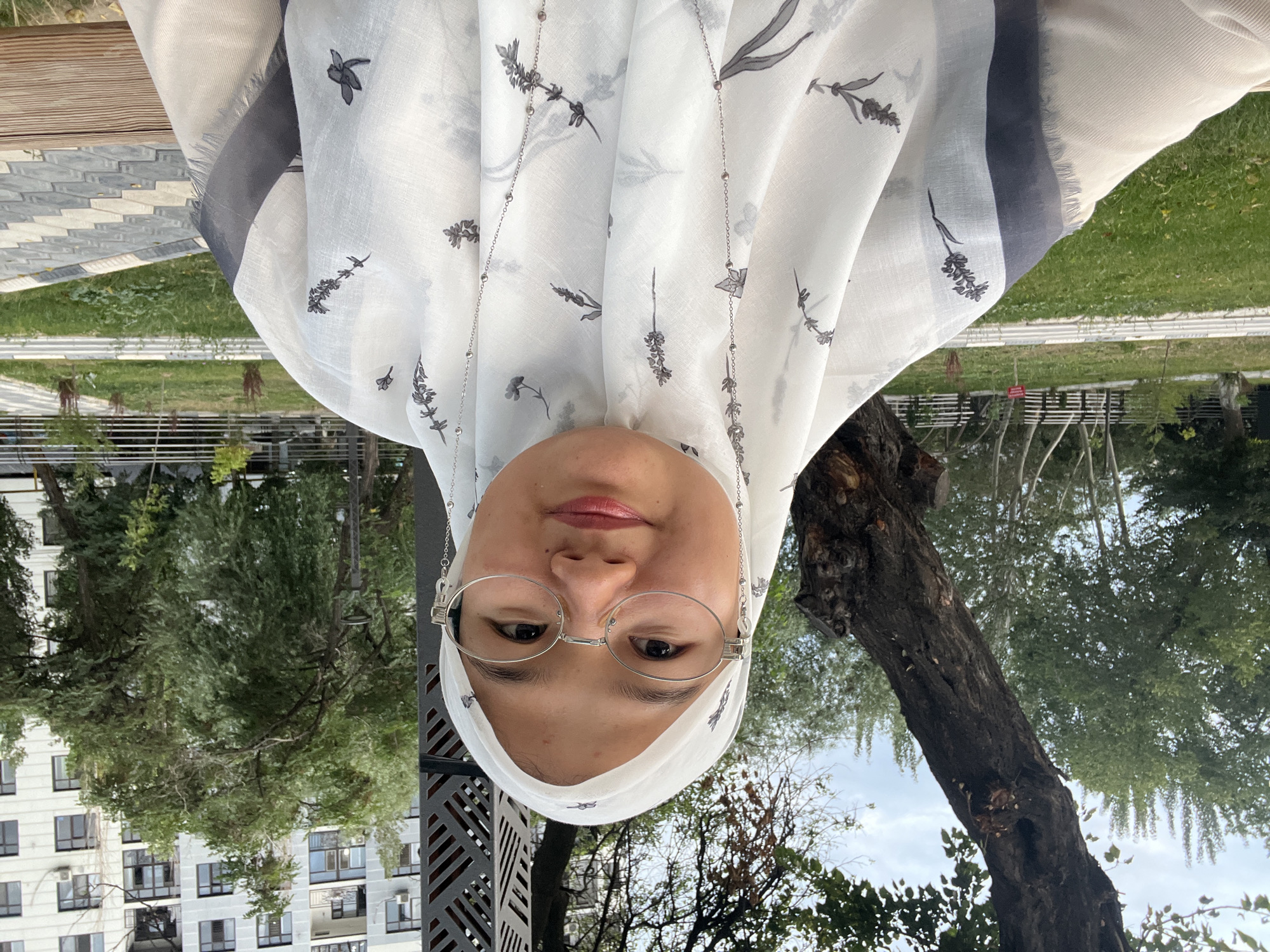 A young woman wearing a white hijab with a floral pattern and silver chain necklace, and clear-rimmed glasses is sitting outdoors on a bench in a park. She is looking directly at the camera and smiling. Behind her, there are trees, a modern building, and a paved walkway. The location appears to be a park in Osh City, Kyrgyzstan, as indicated by the presence of the iconic Sulayman-Too mountain in the background. The environment is green and lush, suggesting a pleasant day.
