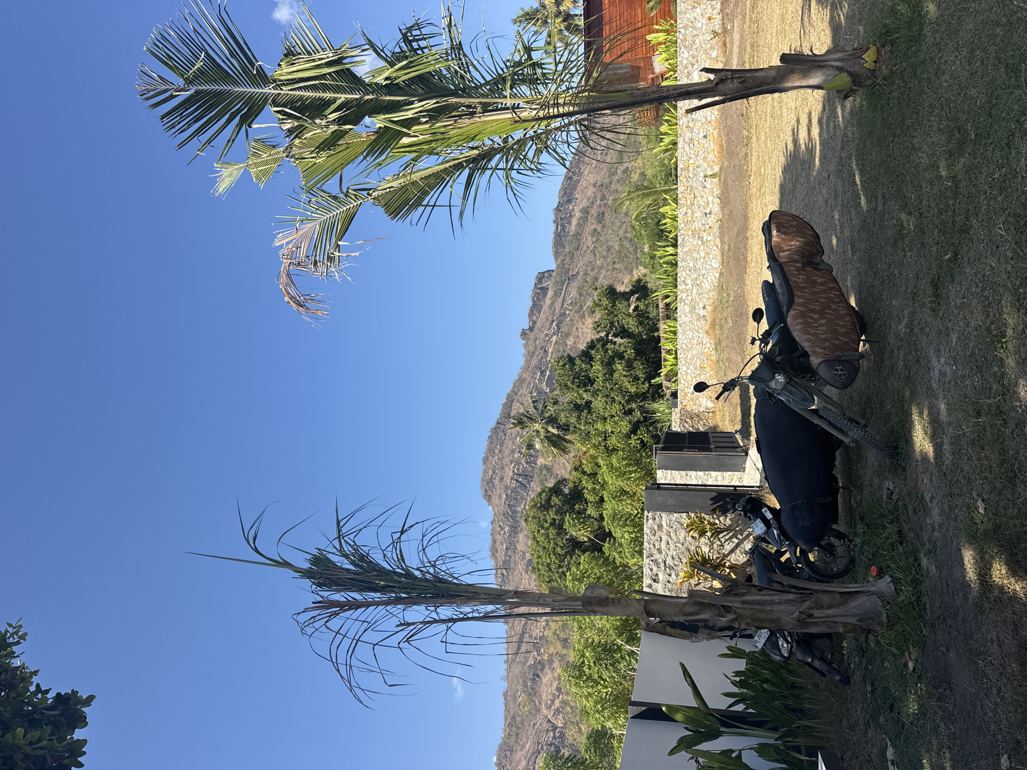 This is a medium shot of an outdoor scene on a clear day, likely in Indonesia. In the foreground, two motorcycles are parked on a grassy area. The motorcycle on the right has a sidecar with a camouflage cover. Behind the motorcycles, there's a low stone wall, and beyond that, lush green trees and vegetation. In the background, a dry, rocky mountain range rises against a bright blue sky. There is a wooden building on the right side of the image. The scene is peaceful and natural.