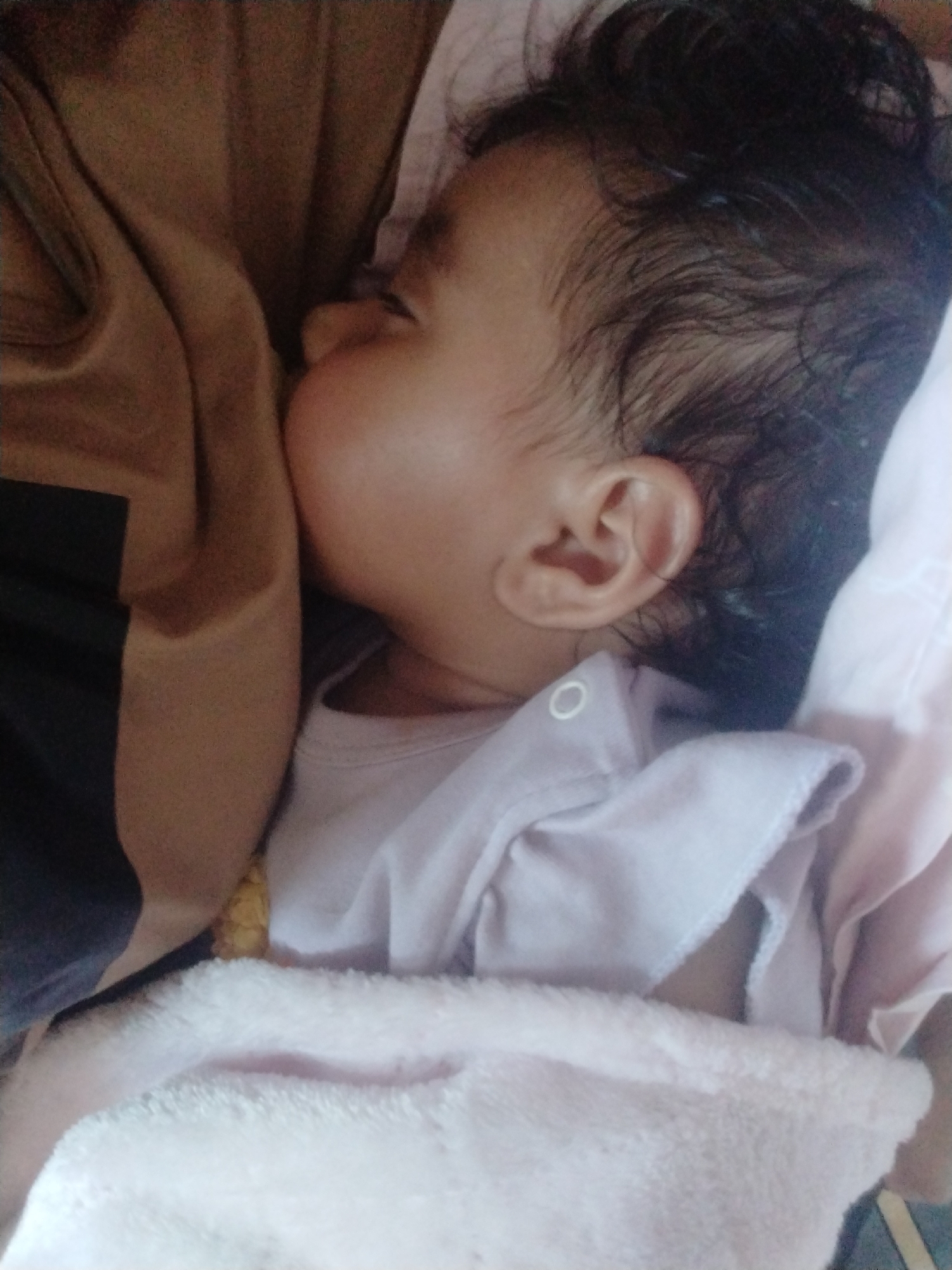 A baby is breastfeeding, lying on their side with their face turned towards a person's chest. The baby is wearing a light-colored shirt and is partially covered with a soft, pink blanket. The person breastfeeding the baby is wearing a tan shirt with a black sleeve visible on the left side of the frame. The baby has dark, curly hair. The location is Unknown, Unknown.