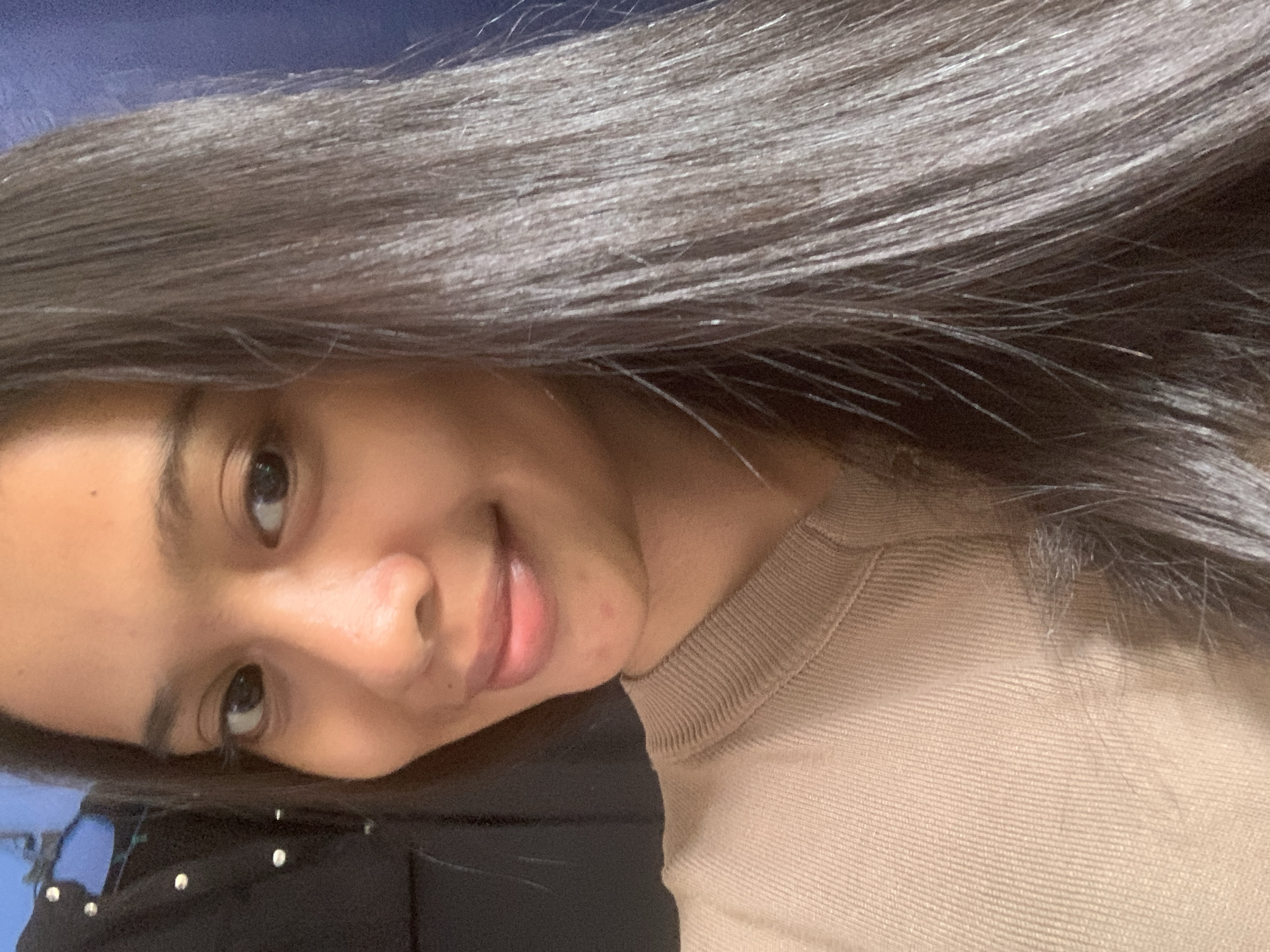 The image is a close-up selfie of a young woman with long, dark hair. She is wearing a tan-colored, ribbed crew neck sweater. The background is a blurred dark blue curtain on the right and a dark object, possibly clothing, on the left with some visible white buttons. The woman's face is angled slightly to the left, and she is smiling. Her skin appears clear with some subtle redness on her chin. The lighting suggests an indoor setting. The image does not contain any information indicating its location in Sungai Meriam, Indonesia.