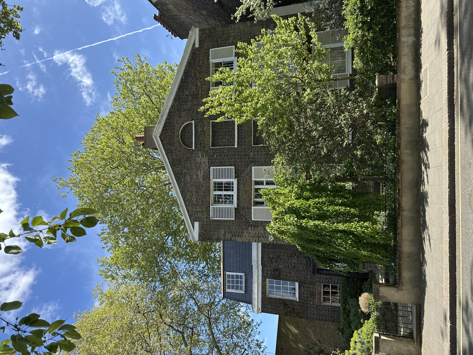 An outdoor scene captures a multi-story brick house on a bright, sunny day, viewed from a street-level perspective looking upwards. The dominant structure is a large, dark reddish-brown brick house, characterized by numerous white-framed windows, many of which feature white exterior shutters or blinds. The upper part of the house presents a steeply pitched gable with a distinctive white trim and a semi-circular arched opening, flanked by rectangular windows. Below this, a section with a dark grey slate roof and additional windows protrudes. Green ivy or climbing plants are visible on the lower brickwork.
Lush, bright green trees with dense foliage frame the house, particularly on the left side, with some branches extending into the foreground. Above the house and trees, a clear blue sky is visible, adorned with scattered white clouds and a faint airplane contrail stretching across.
To the right of the house, a paved sidewalk or street runs parallel to the property, featuring visible curb lines and square paving slabs. Shadows from trees and the building are cast across the pavement, indicating strong sunlight. Black metal railings or fences define the property boundary.
The scene depicts a quiet residential area, consistent with an urban or suburban setting, and the architectural style suggests London, United Kingdom. There are no people visible, nor any discernible text. The strong sunlight and clear shadows suggest it is daytime, likely late morning or early afternoon.