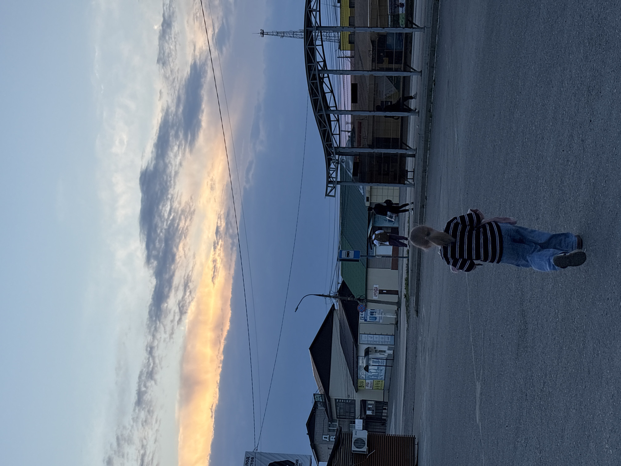 The image shows a street scene in Chaikovsky, Russia. The sky is predominantly blue with bands of white and yellow clouds on the left side. An individual is walking along the paved road, wearing blue jeans, a black and white striped shirt, and grey footwear. There are buildings lining the road, and a bus stop can be seen with people waiting under it. The buildings have multiple signs and details in Russian. A metal structure, possibly a shelter or framework, is on the upper right side. Power lines are visible stretching across the sky.