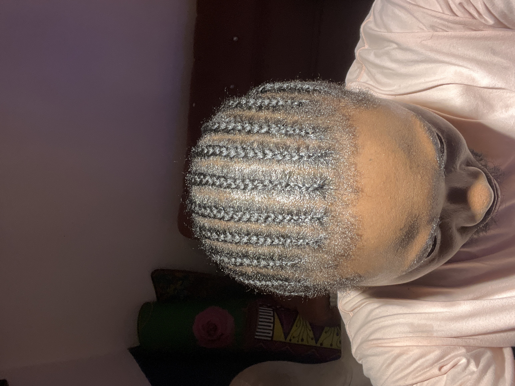 A person is shown from the shoulders up, with their head bowed. The individual has dark skin and is wearing a light pink t-shirt. Their hair is styled in neat, thin braids that run from the front of their head towards the back. The background is dimly lit and appears to be an indoor setting, with a dark wall and a hint of furniture or a decorated surface to the left. The lighting suggests a single light source from above, casting shadows and highlighting the braids. The location is identified as the city of Jos, Nigeria.