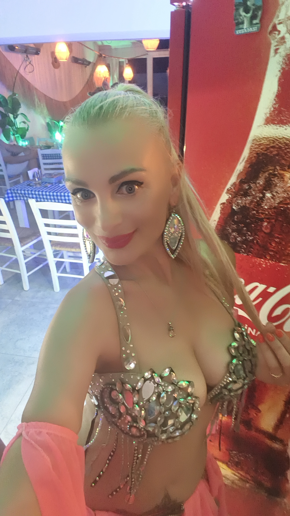 A blonde woman, wearing a bejeweled belly dancing costume with pink flowing sleeves, poses for a selfie. She has dark eye makeup and bright red lipstick. She is standing in front of a large, red Coca-Cola advertisement, possibly at an outdoor restaurant or bar in Kuşadası, Turkey. In the background, there are tables with blue and white checkered tablecloths and white chairs. String lights are visible overhead, and green plants add to the ambiance. The lighting suggests it is evening or nighttime.