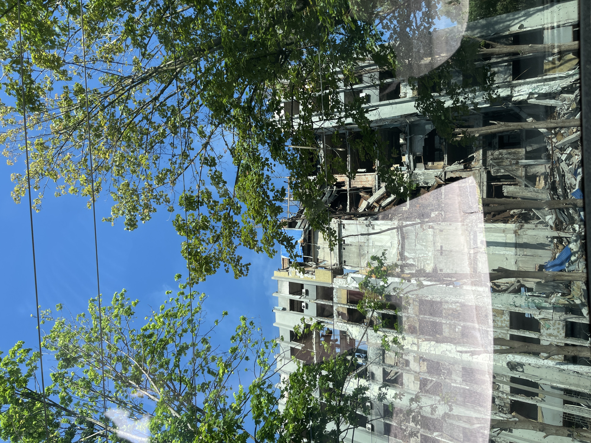 The image, oriented vertically and possibly taken from inside a vehicle due to prominent reflections, displays multi-story buildings suffering severe damage, viewed through the vibrant green canopy of several trees against a bright blue sky.
The buildings appear to be extensively destroyed, with numerous windows missing, revealing gutted interiors filled with debris, rubble, and shattered construction materials. Facades are crumbling, with plaster peeling off and structural elements exposed. On one of the buildings, several air conditioning units are still visible, alongside a blue tarp covering a section of its upper structure or roof. Large broken branches or wooden beams are strewn within the damaged sections of the buildings.
The scene is bathed in bright daylight under a clear, sunny sky. Reflections on what appears to be a vehicle window are visible across the image, distorting parts of the trees and buildings. Thin utility lines are also discernible against the sky, both directly and in the reflections.
No specific text is visible within the scene. The extensive destruction of urban infrastructure, coupled with the location context of Dnipro, Ukraine, strongly suggests the aftermath of conflict or shelling. The image captures a stark contrast between resilient nature and man-made devastation.