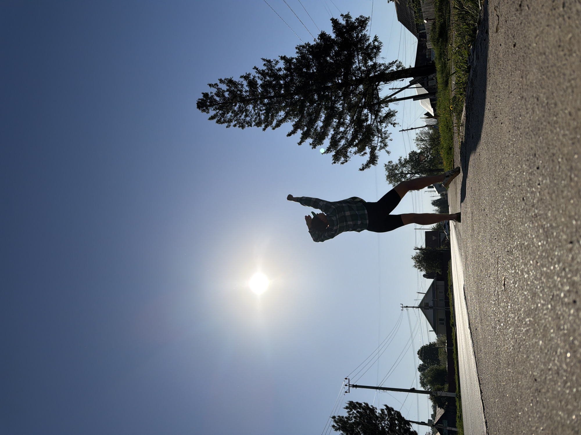 A male figure wearing a plaid shirt and dark shorts is silhouetted against the bright sun, appearing to jump in the air on a paved road. The perspective is low, looking upwards, with the clear blue sky filling the majority of the frame. Power lines stretch across the sky. A tall conifer tree and several houses are visible in the background, along the edge of the road. The location is likely a residential street in the city of Chaikovsky, Russia. The overall impression is that the image was taken at mid-day, given the position of the sun.