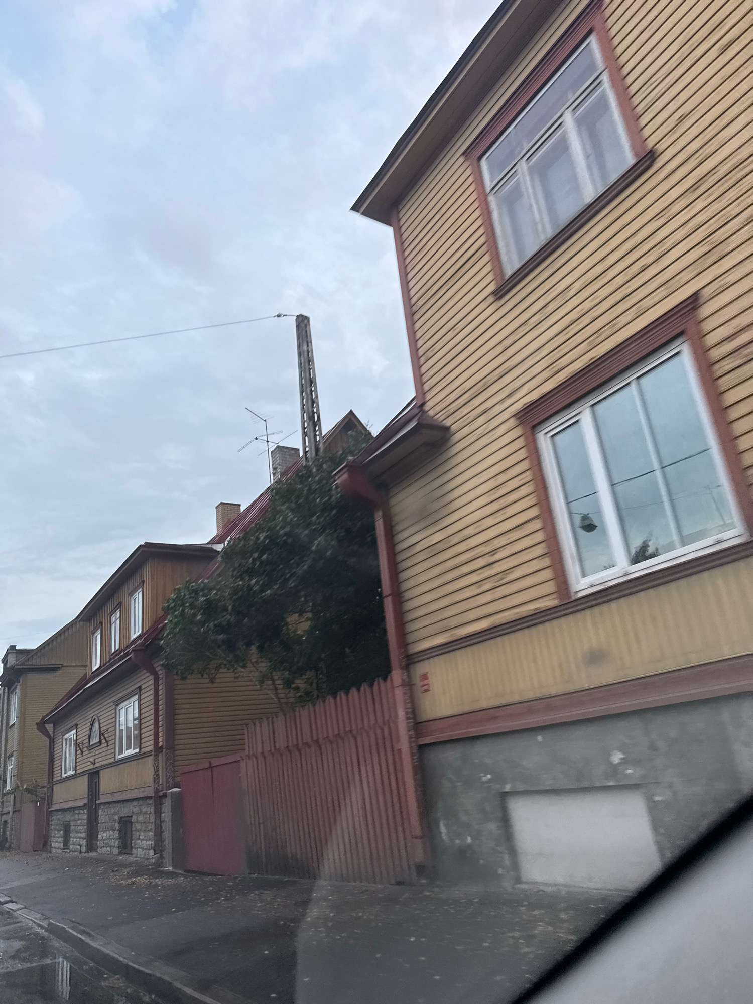 A medium-shot, tilted perspective captures a row of traditional wooden houses along a street in Tallinn, Estonia. The houses are painted in muted yellow and brown tones with red trim around the windows and roofs. The buildings are of varying heights and architectural styles, with some featuring multiple stories and visible dormer windows. A red wooden fence separates the properties from the street. The sky is overcast with soft gray clouds. A tall, thin pole with antenna-like structures rises behind one of the houses. The street is wet, suggesting recent rain, and fallen leaves are scattered on the asphalt and sidewalk. A portion of a car's windshield is visible in the foreground, indicating the image was taken from inside a vehicle.