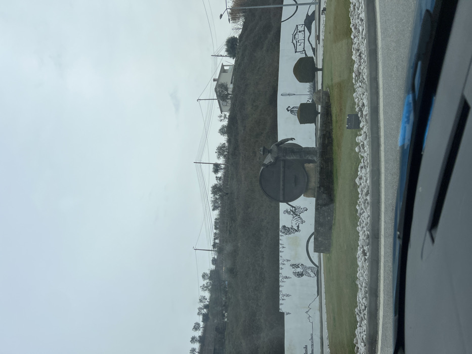 The image displays a roundabout in Santa Marta de Penaguião, Portugal, viewed from inside a vehicle. In the center of the roundabout, a sculpture depicts a male figure wearing a hat, standing alongside a large, dark-colored barrel positioned on a stone base. Behind this monument, a curved white wall features black line-art murals, illustrating grapevines, a person harvesting grapes, and the outline of a building and another figure. The central island of the roundabout is covered in green grass, bordered by a ring of light-colored rocks, and includes two dark, trimmed hedges to the right of the main sculpture.
A steep, sparsely vegetated hill dominates the background, with scattered small trees visible across its surface. A white house with a dark roof stands on the upper portion of the hill to the right. Utility poles and multiple power lines span horizontally across the mid-ground. The sky is overcast and uniformly light grey. The bottom foreground shows the top edge of a car's dashboard and windshield, with a blue reflection or object visible on the dash.