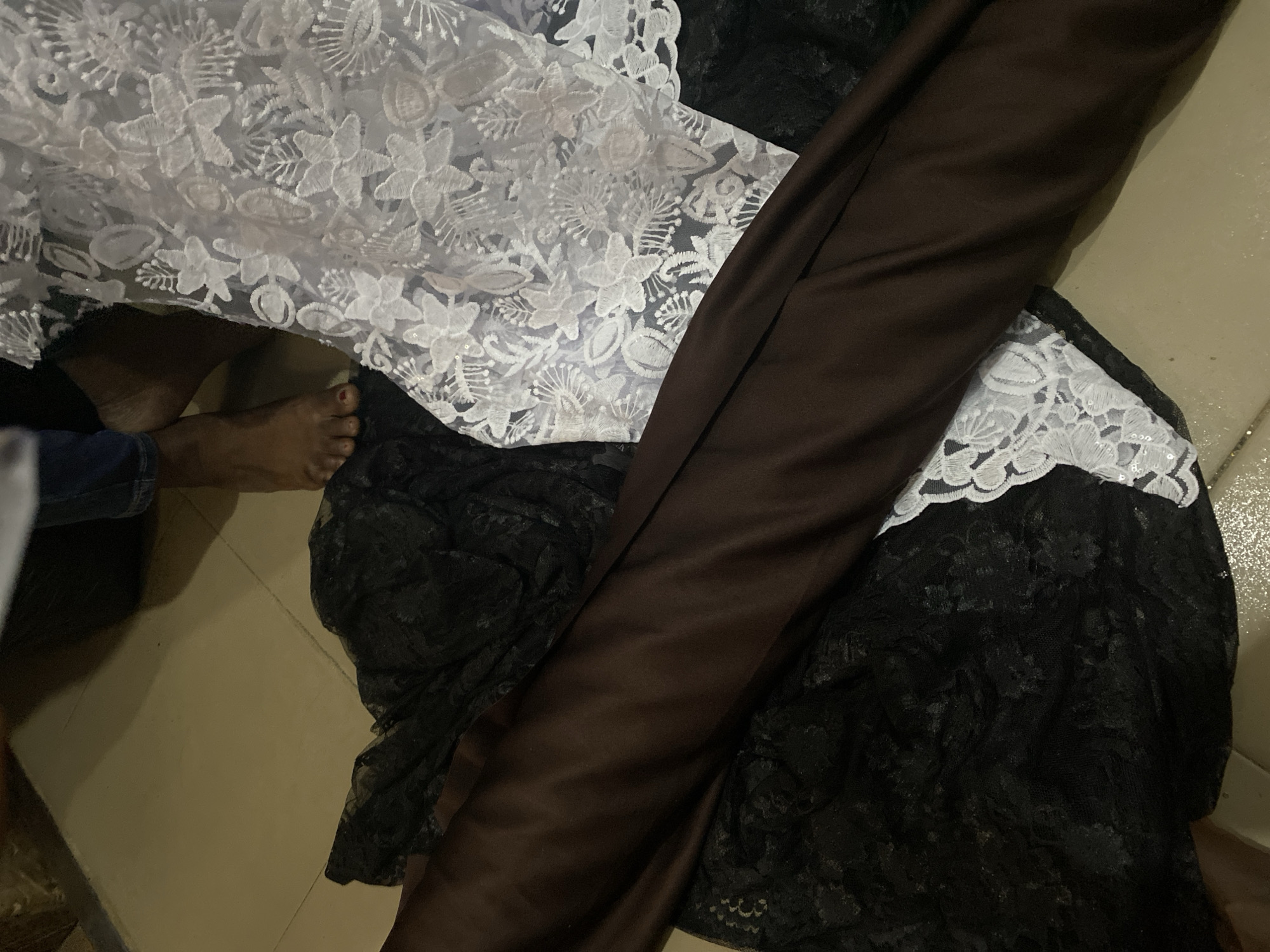 The image depicts a close-up, overhead view of several fabrics, including a large piece of dark brown satin, a black lace fabric with floral patterns, and a white lace fabric also with intricate floral designs. A person's bare feet, with painted toenails, are visible in the upper left portion of the frame, along with the bottom of their jeans. The fabrics are spread out on a tiled floor, which appears to be in a residential setting within the city of Jalingo, Nigeria. The lighting is dim, creating shadows and highlighting the textures of the materials.