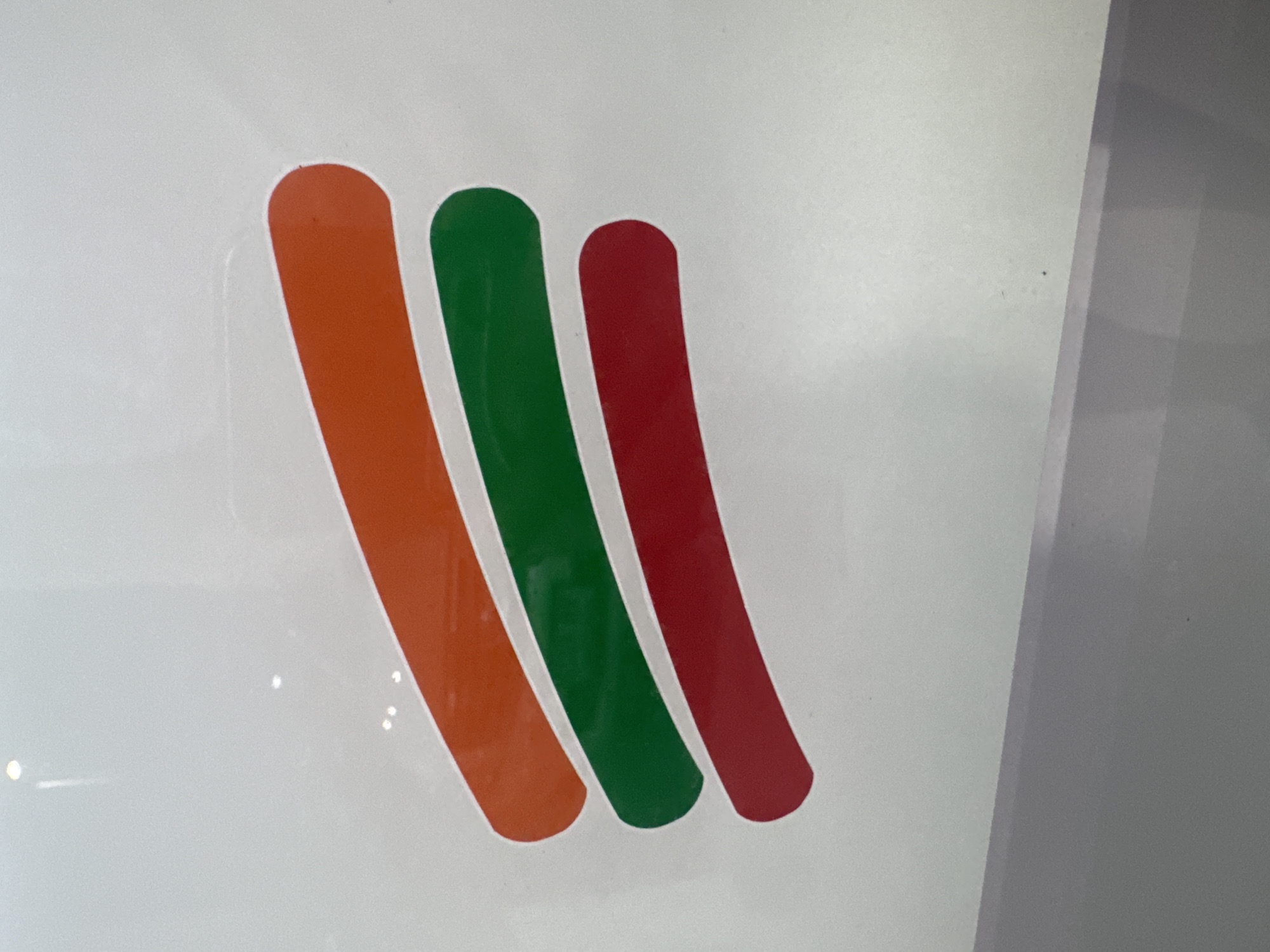 The image displays a close-up of a partial logo consisting of three horizontally aligned, slightly curved, and rounded-edge bars, stacked vertically on a light gray, reflective surface. The top bar is orange, the middle bar is green, and the bottom bar is red. Multiple small, bright specular reflections are visible on the glossy background surface, suggesting a polished or glazed material. No discernible text, individuals, or additional contextual elements are present. Based on the distinct color sequence and design, which strongly resembles the stylized "7" found in the branding of 7-Eleven convenience stores, this image is identified as a detail from a 7-Eleven outlet. Given the extreme prevalence and ubiquity of 7-Eleven stores throughout the city, the location of this scene is situated in Bangkok, Thailand.