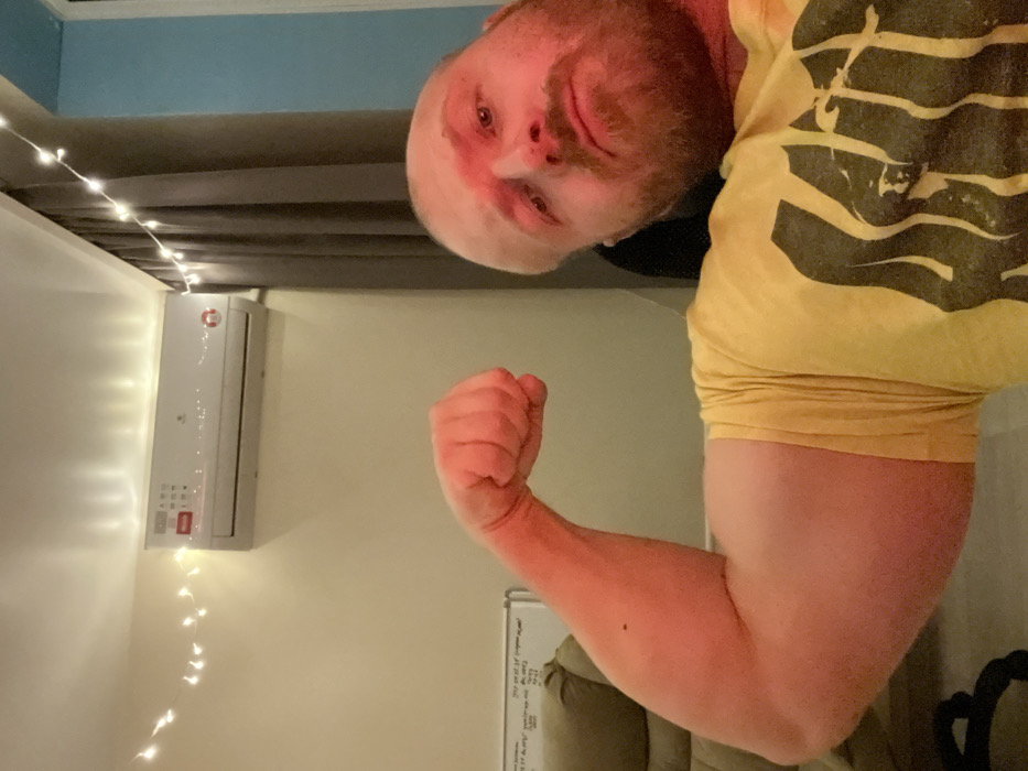 A person is posing while facing the camera, with their bicep flexed. They are wearing a yellow t-shirt. Behind them is a wall-mounted air conditioner, surrounded by small string lights. A portion of a curtain and the ceiling are visible at the top. A whiteboard is partially visible in the lower portion of the image. The picture is slightly rotated.