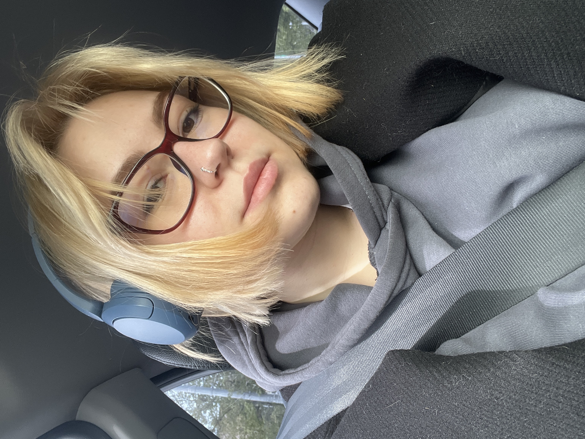 A selfie taken inside a car. A young woman with blonde shoulder-length hair, wearing reddish-brown glasses and a silver nose ring, is the main subject. She is wearing a gray hooded sweatshirt and a black coat. A blue headphone rests behind her ear. The car's interior is visible, including the seatbelt and dashboard. The background shows a glimpse of greenery through the car window. The image doesn't provide sufficient information to identify the exact location in Odesa, Ukraine.
