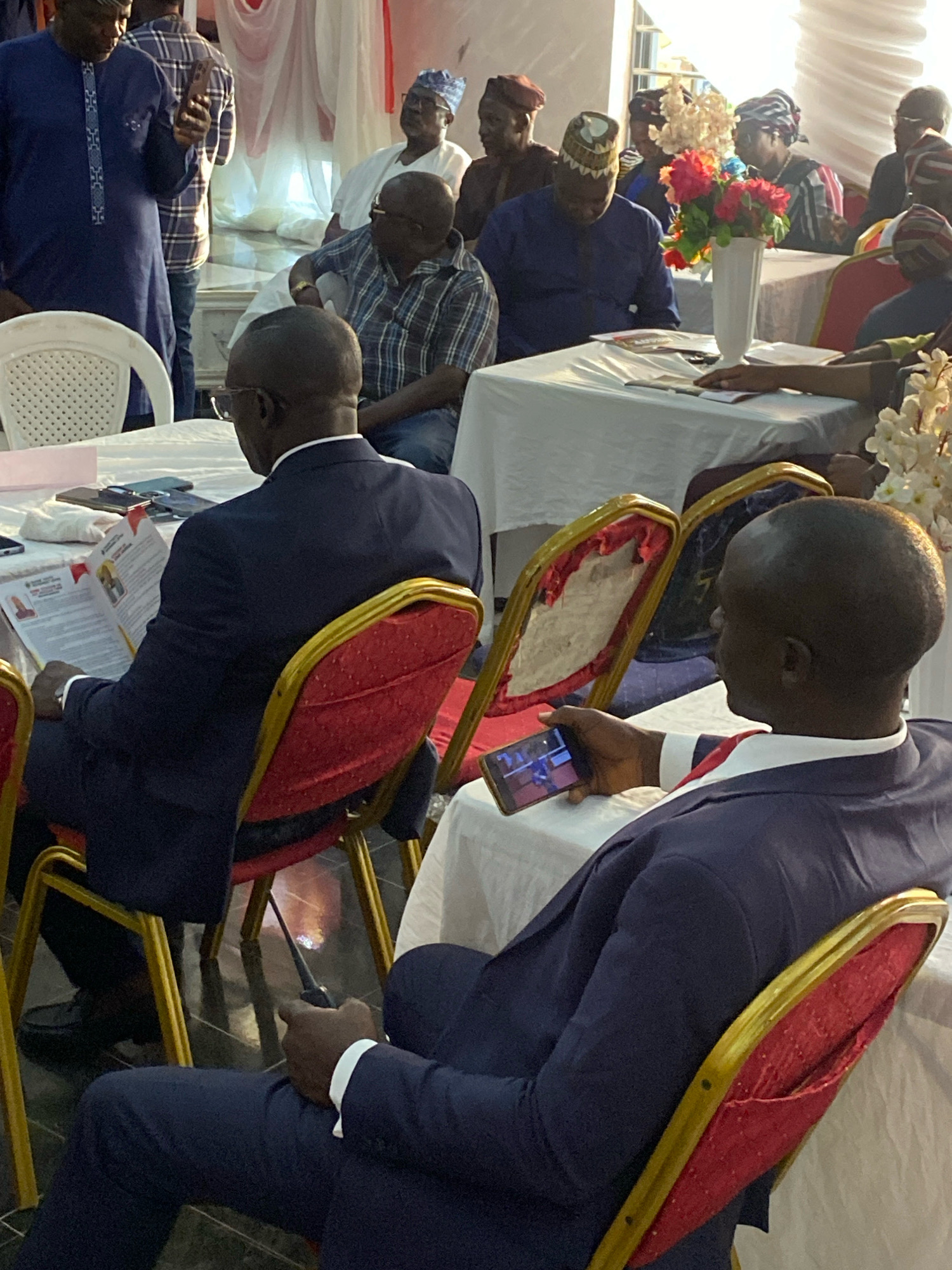 The image depicts an indoor gathering in Jos, Nigeria. Several individuals, primarily men, are seated at tables draped with white cloths, suggesting a formal event like a conference, seminar, or meeting. Some attendees are engaged with documents, while others are using their mobile phones. One person in the foreground is engrossed in reading a document, while another is looking at their phone. The environment appears to be decorated with white drapes and artificial flowers, indicating a celebratory or ceremonial occasion. The lighting suggests it is daytime, possibly in a hall or event center.