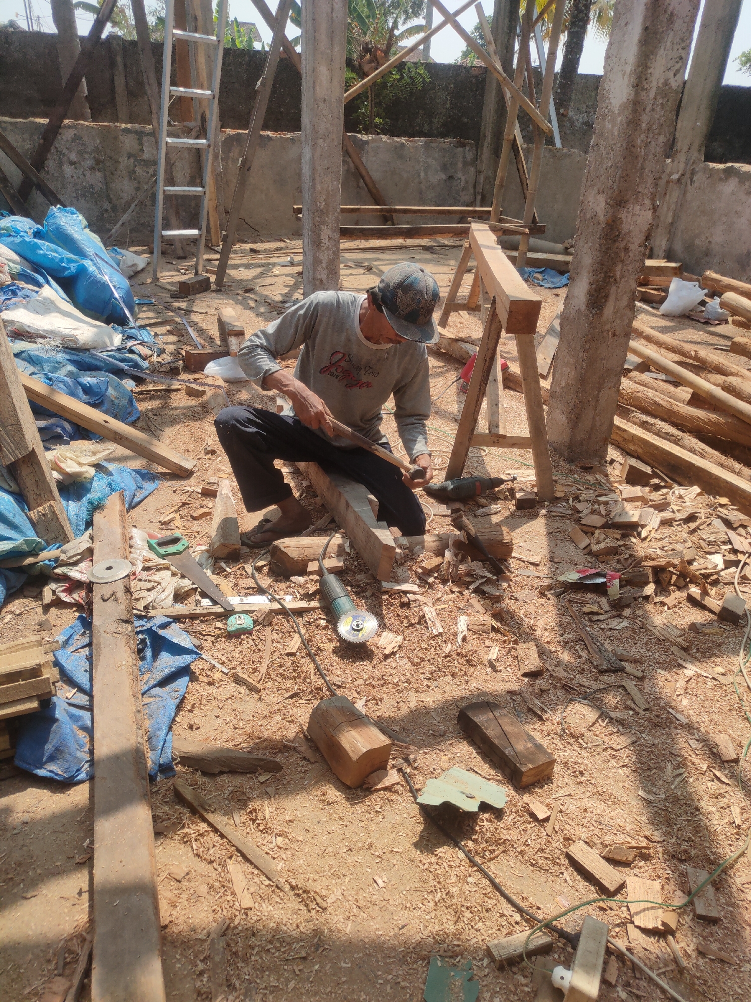 This image captures a construction site in Sumedang, Indonesia, where a man is actively working with wood. He is dressed in a grey long-sleeved shirt, dark pants, and a cap, sitting on the ground amidst sawdust and wood debris. A power grinder is positioned near him, and he appears to be using a hand tool on a piece of wood. The environment suggests an unfinished building, with wooden scaffolding, concrete pillars, and building materials such as blue tarps and piles of lumber scattered around. The ground is covered in wood shavings and fragments, indicating recent woodworking activity. A ladder is visible in the background, leaning against a wall. The scene is outdoors, under daylight, with shadows indicating the time of day.