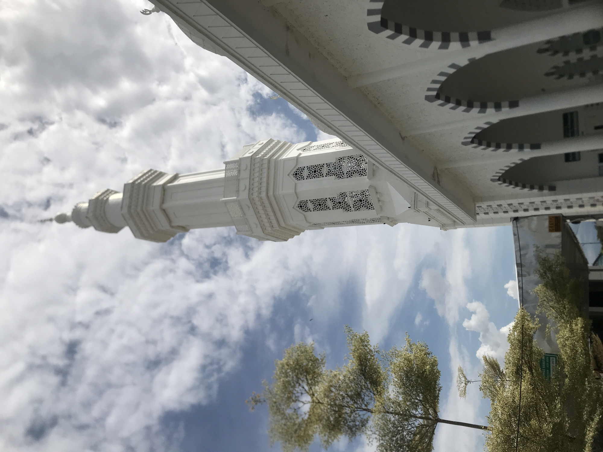 The image shows a white minaret against a cloudy sky, partially framed by a white building structure with circular decorations. Trees are visible in the foreground. The location is inferred to be Meunasahaluedrienlhoksukon Barat, Indonesia. The image is taken from a low angle, looking upwards, with the camera oriented upside down.
