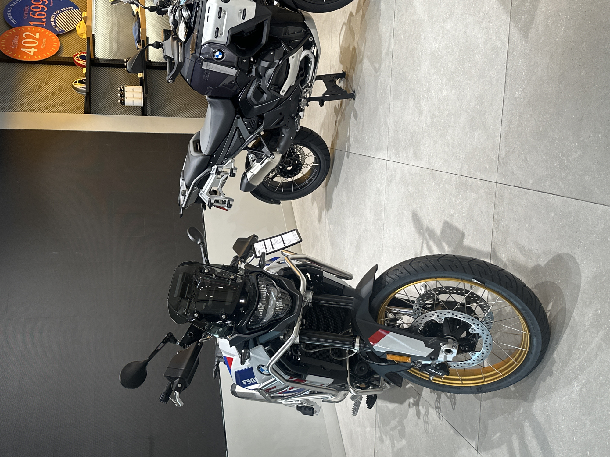 This indoor scene features two motorcycles on display. The first, closer to the camera, is a white, blue, and red BMW F900 GS Adventure motorcycle. Its front wheel has a gold rim and Brembo brakes. A sign on the motorcycle reads "F90" and "Brembo". Behind the F900 GS, there is a black BMW R 1250 GS motorcycle. The floor is gray tile. In the background, against a honeycomb wall, are circular signs reading "402" and "1.699" and the phrase "NICE TO MEET YOU AGAIN". Based on the Thai writing on the sign with the number "402", this image was likely taken in Bangkok, Thailand.