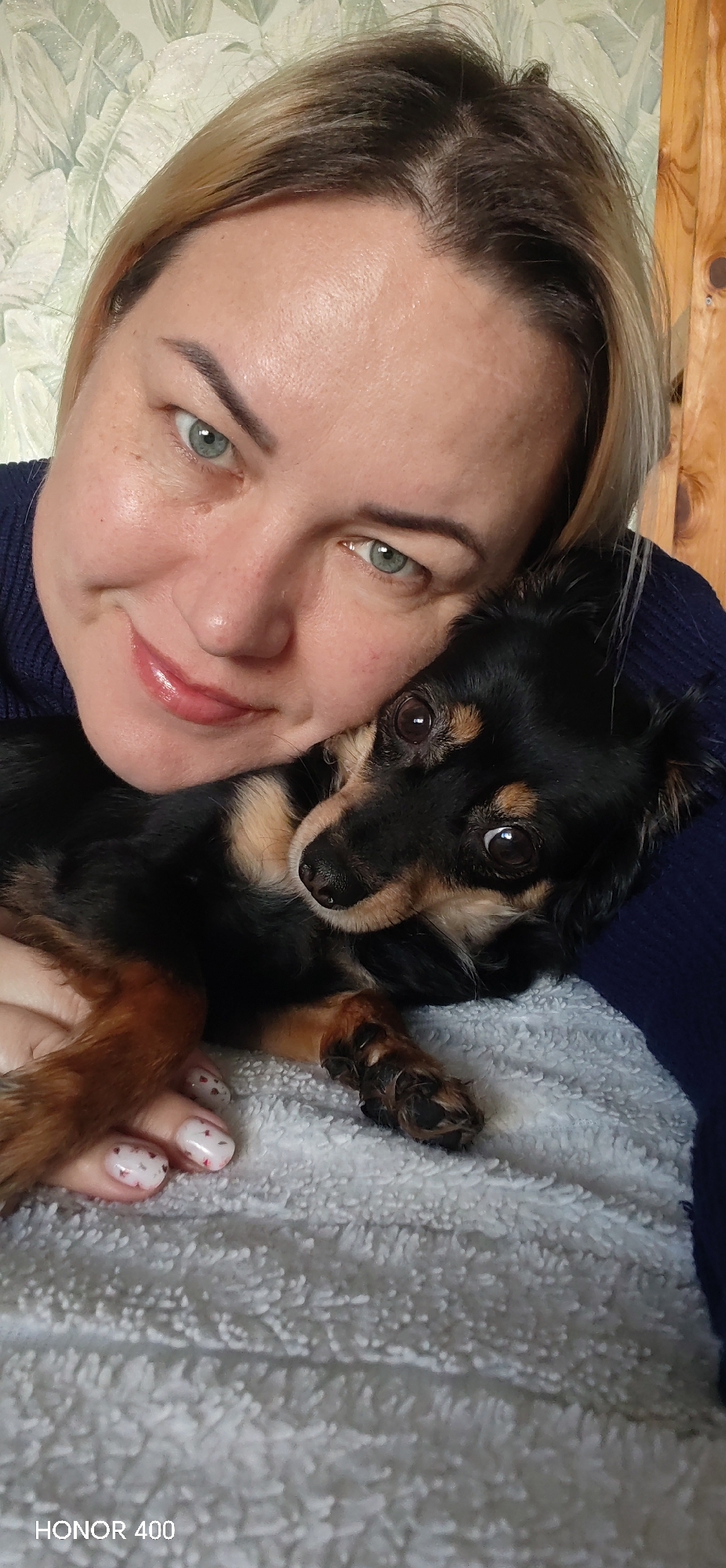 A woman with blonde hair and green eyes is holding a small black and tan dog against a soft, white blanket. The dog is looking at the camera. The woman is smiling. The background appears to be a floral wallpaper. The image includes "HONOR 400" in the lower-left corner. It is unclear if the location is in Noviy, Russia.