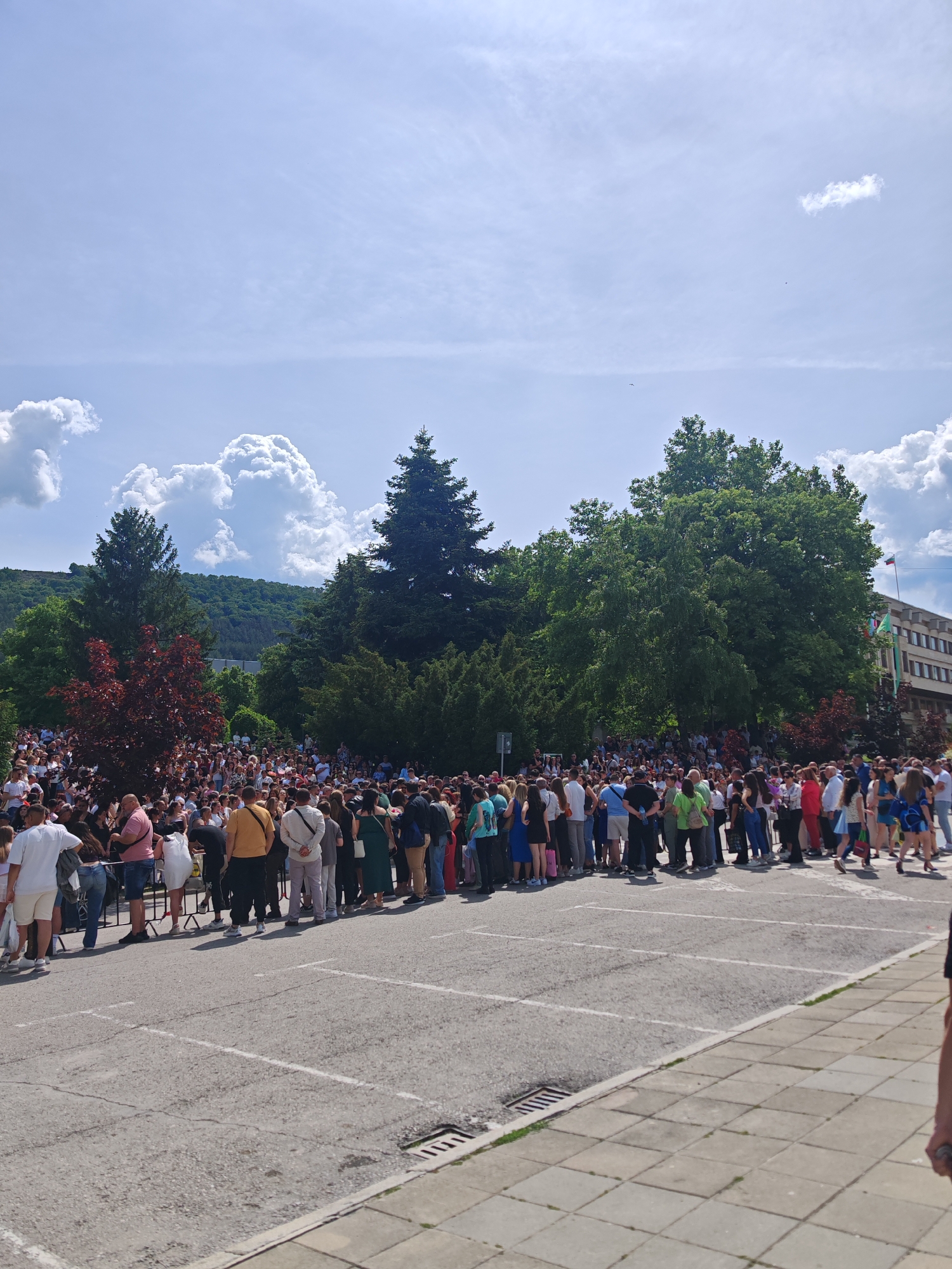 A large crowd of people is gathered in front of a building with a Bulgarian flag. The crowd appears to be waiting in line. The location is likely the city of Shumen, Bulgaria. The environment includes trees and a cloudy sky. The surface in front of the crowd is paved with gray parking lines.