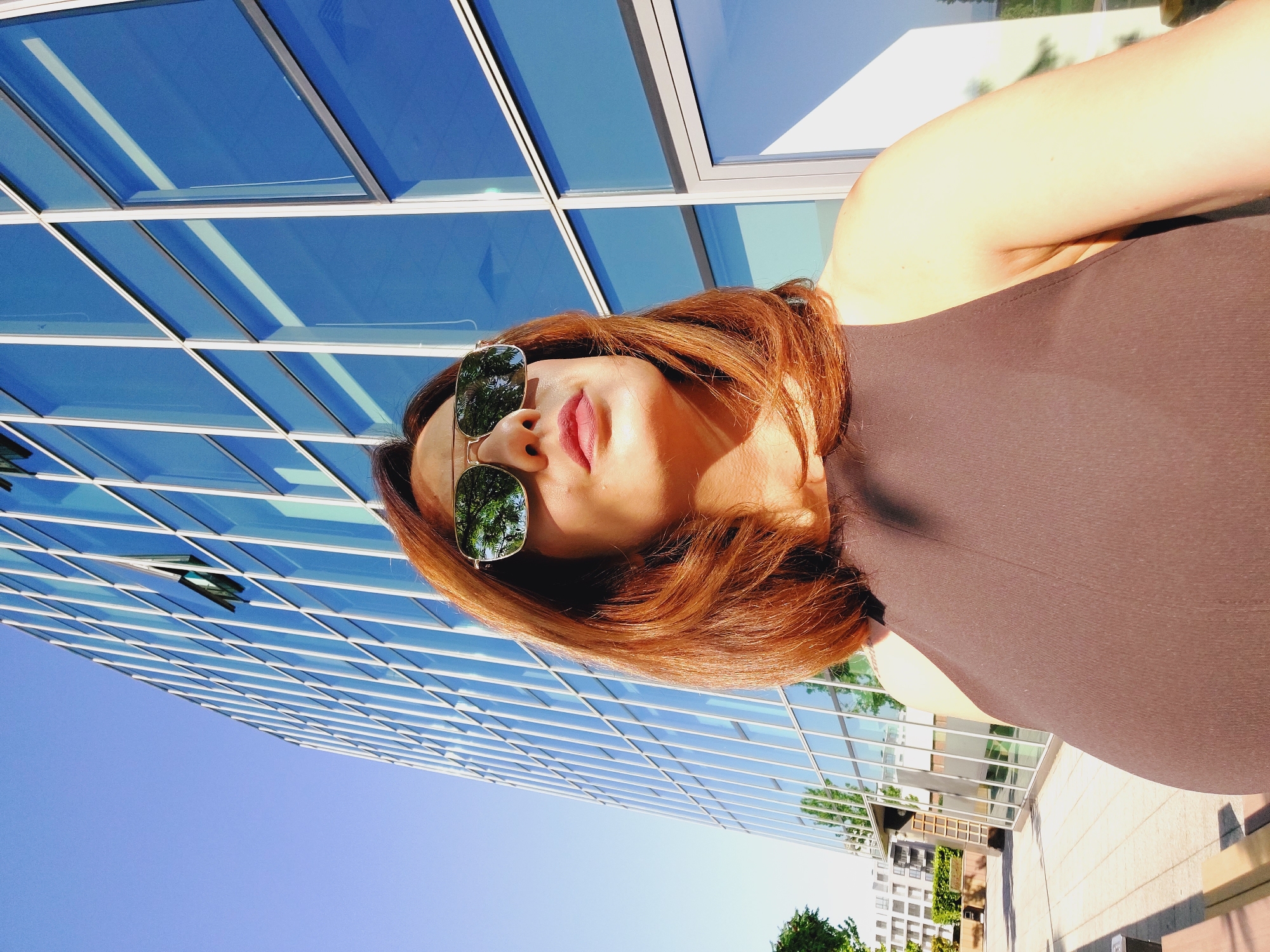 In Sofia, Bulgaria, a woman with auburn hair and sunglasses looks upward, reflecting a tree-lined sky in her lenses. She is positioned in front of a modern glass-faced building, its windows angled to catch the bright sunlight. The woman is wearing a dark, sleeveless top. The background features a clear blue sky and the architectural lines of the building, creating a sense of urban elegance.