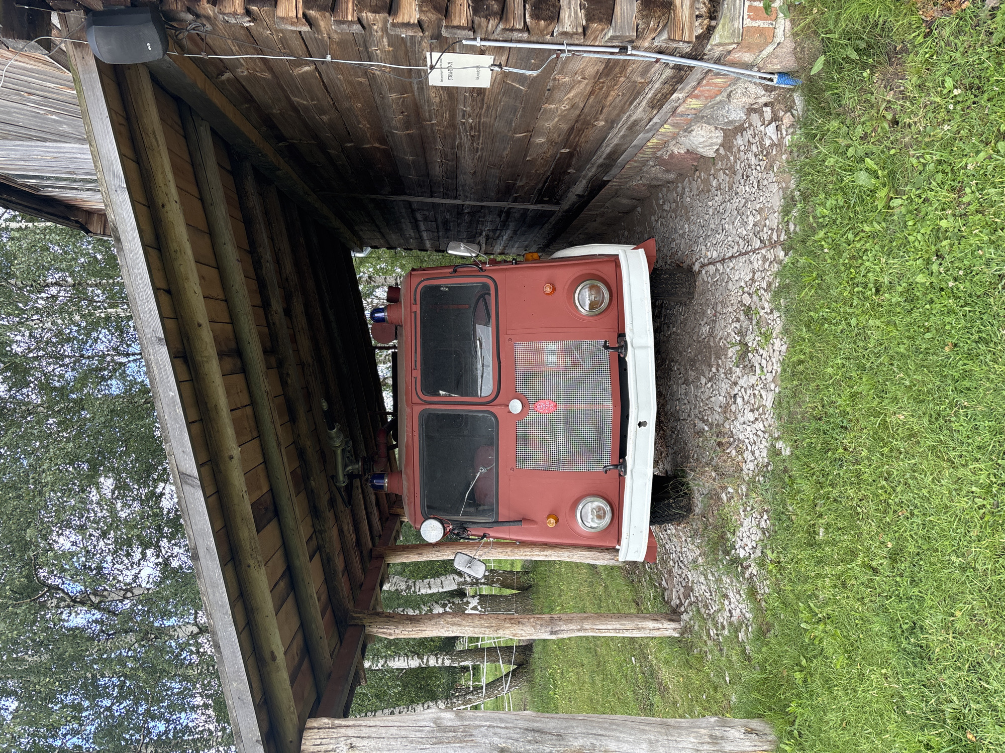 A red vintage fire truck is parked under a wooden shelter next to a rustic wooden building. The truck is positioned facing forward, with its headlights, grille, and red emergency lights visible. The shelter's roof is constructed of wooden planks, supported by rough-hewn wooden posts. To the right of the truck, the wooden building features weathered planks and a window. A white sign with text is affixed to the building's exterior. The ground in front of the truck is covered with gravel and stones, while the surrounding area is overgrown with green grass. In the background, trees with green foliage are visible. The scene appears to be located in Sielachowskie, Poland.