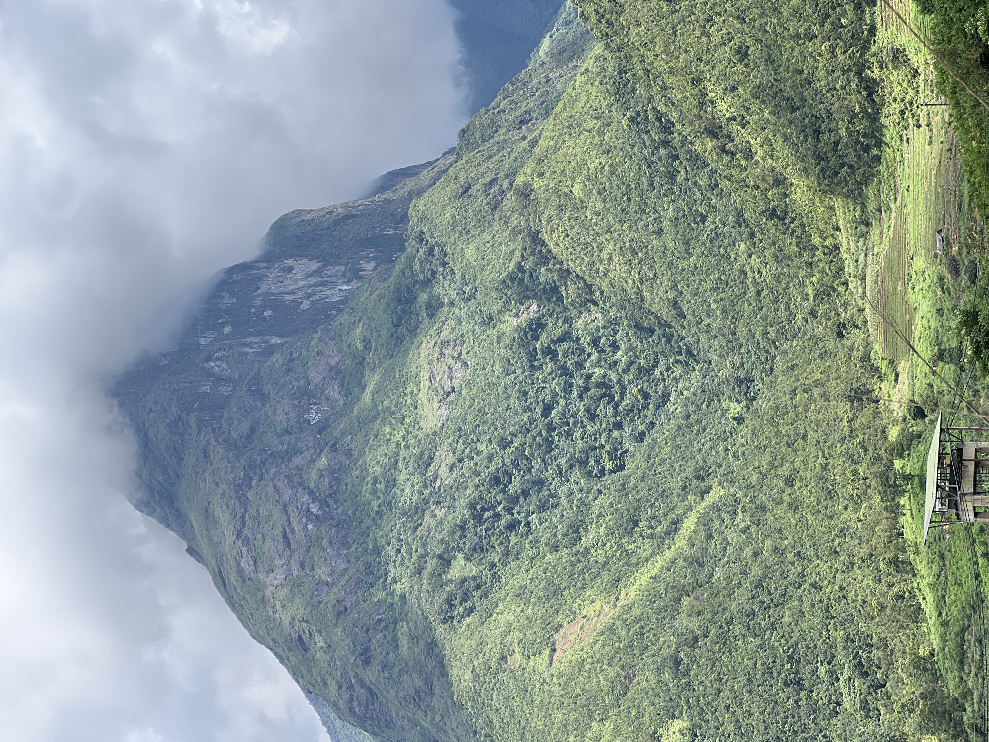 The image captures a dramatic mountainous landscape near the city of Bình Lư, Vietnam. The foreground features a man-made structure, possibly an observation deck or a small building, elevated on concrete stilts. This structure is situated on a verdant, gently sloping hill. Beyond this, the terrain rises sharply, covered in dense, lush green vegetation that blankets the steep mountainsides. The upper reaches of the mountains are partially obscured by thick, grey clouds, lending a dramatic and somewhat mysterious atmosphere to the scene. The overall impression is one of untamed natural beauty, with a touch of human intervention visible in the foreground.
