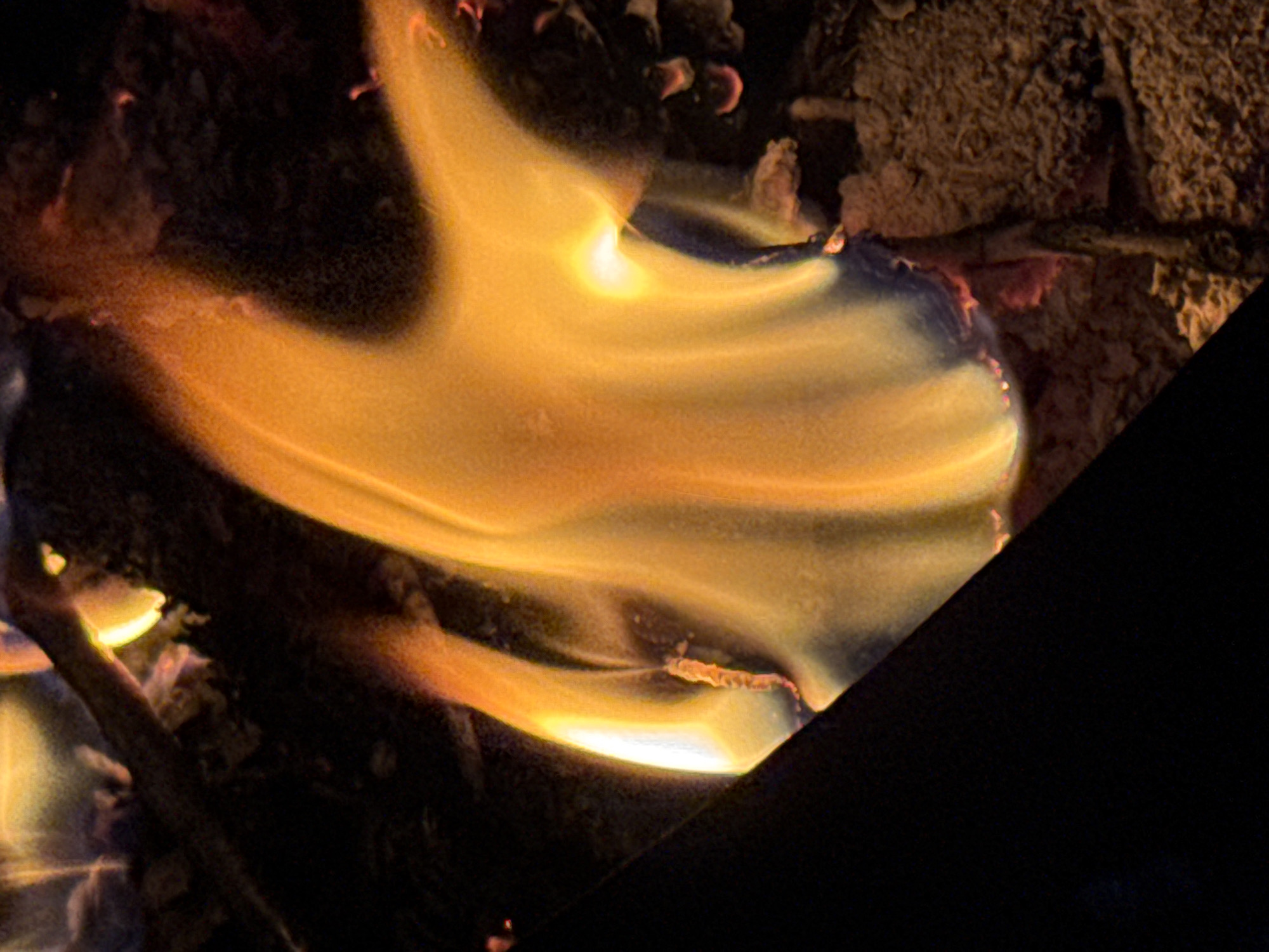 The image displays a close-up view of a fire, with flames in shades of orange, yellow, and white. The fire is consuming what appears to be wood or paper, indicated by charred edges and a dark, textured background. The lighting is dim, with the fire being the primary light source, casting a warm glow. A dark, diagonal object, possibly a piece of metal or stone, cuts across the left side of the frame, creating a stark contrast with the bright flames. The overall scene suggests an uncontrolled burn or a bonfire in a natural or outdoor setting. There are no people visible in the image.