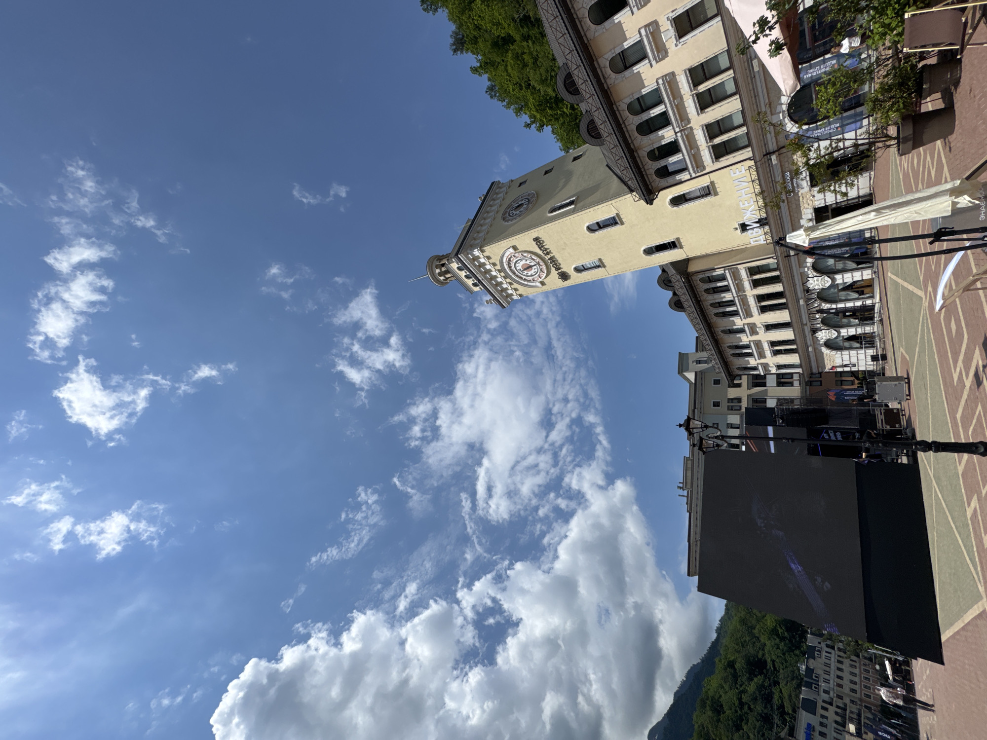 The image shows a wide-angle view of a town square in Estosadok, Russia, on a bright, sunny day with scattered clouds. In the foreground, a large, dark, angled LED screen is set up, suggesting an outdoor event or display. To the right of the screen, a streetlamp stands on a patterned brick pavement. Behind these elements, a cluster of buildings forms the backdrop. Dominating the right side of the frame is a tall, cream-colored clock tower with arched windows, adorned with a clock face that appears to read approximately 1:50. Adjacent to the clock tower is a building with a tiled roof and numerous arched windows. Further in the background, more buildings are visible, some with balconies, and a densely forested hillside rises behind them. Two people are partially visible on the left side of the frame, near the buildings. The sky above is a vibrant blue, with fluffy white clouds.