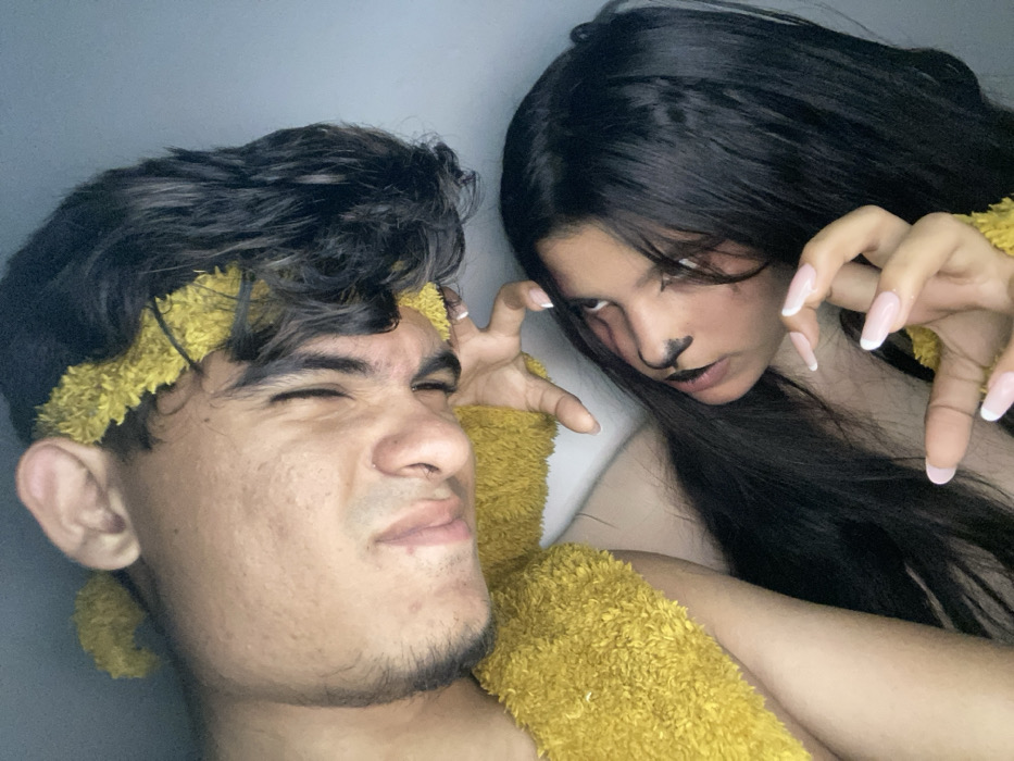 A selfie-style shot shows two people. The individual on the left has dark hair, a yellowish headband, and is pulling a pained expression. The person on the right, slightly behind, has long dark hair, cat-like makeup on their face, and is extending their hands like claws. Both are wearing similar yellow, textured material on their upper arms and torso. The background is a plain, light wall.