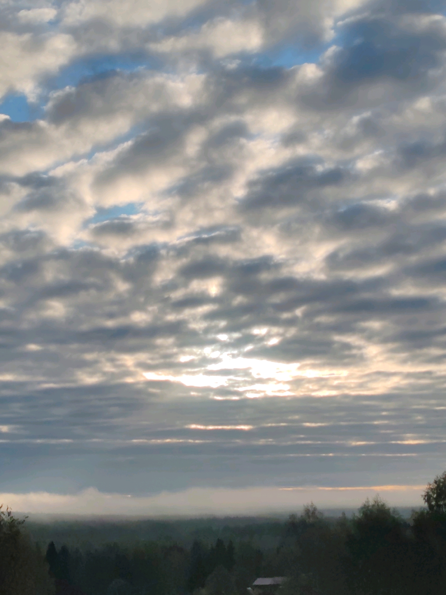 The image captures a cloudy sky over a forested landscape, likely in or near Чухлэм, Russia. The sky is filled with layered clouds, some appearing white and illuminated by sunlight breaking through, while others are darker gray. Patches of blue sky are visible between the clouds. Below the sky, a hazy horizon is discernible, with the silhouettes of trees forming a dense, dark canopy that extends across the frame. A small structure with a white roof, possibly a building or shed, is visible nestled within the trees on the right side of the image, partially obscured by foliage. The overall impression is that of a landscape scene at either sunrise or sunset, given the quality of light filtering through the clouds.