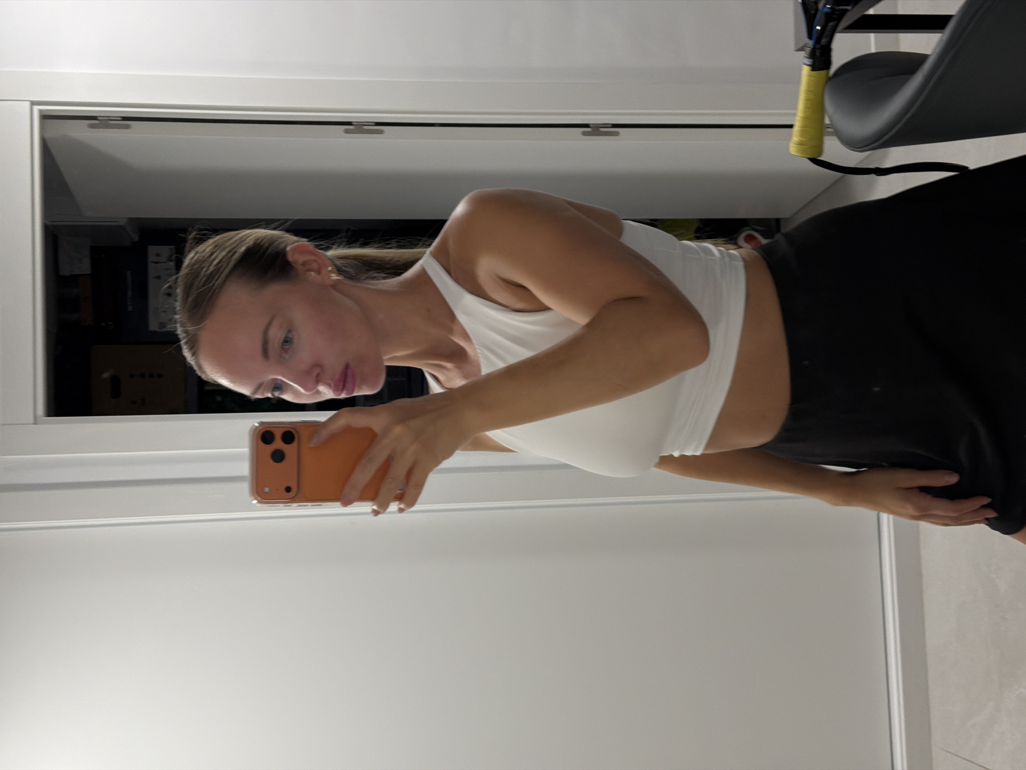 A young Caucasian woman with blonde hair pulled back in a ponytail is taking a mirror selfie indoors. She is wearing a white crop top and a black skirt. She is holding an orange smartphone in her right hand, angled to capture her reflection. In the background, a doorway opens to a room with shelves containing various items. To the right of the doorway, a partial view of a gray chair with a yellow handle is visible. The room appears to be a bedroom or dressing area. The image does not provide any specific visual cues to indicate the location within Finestrat, Spain.