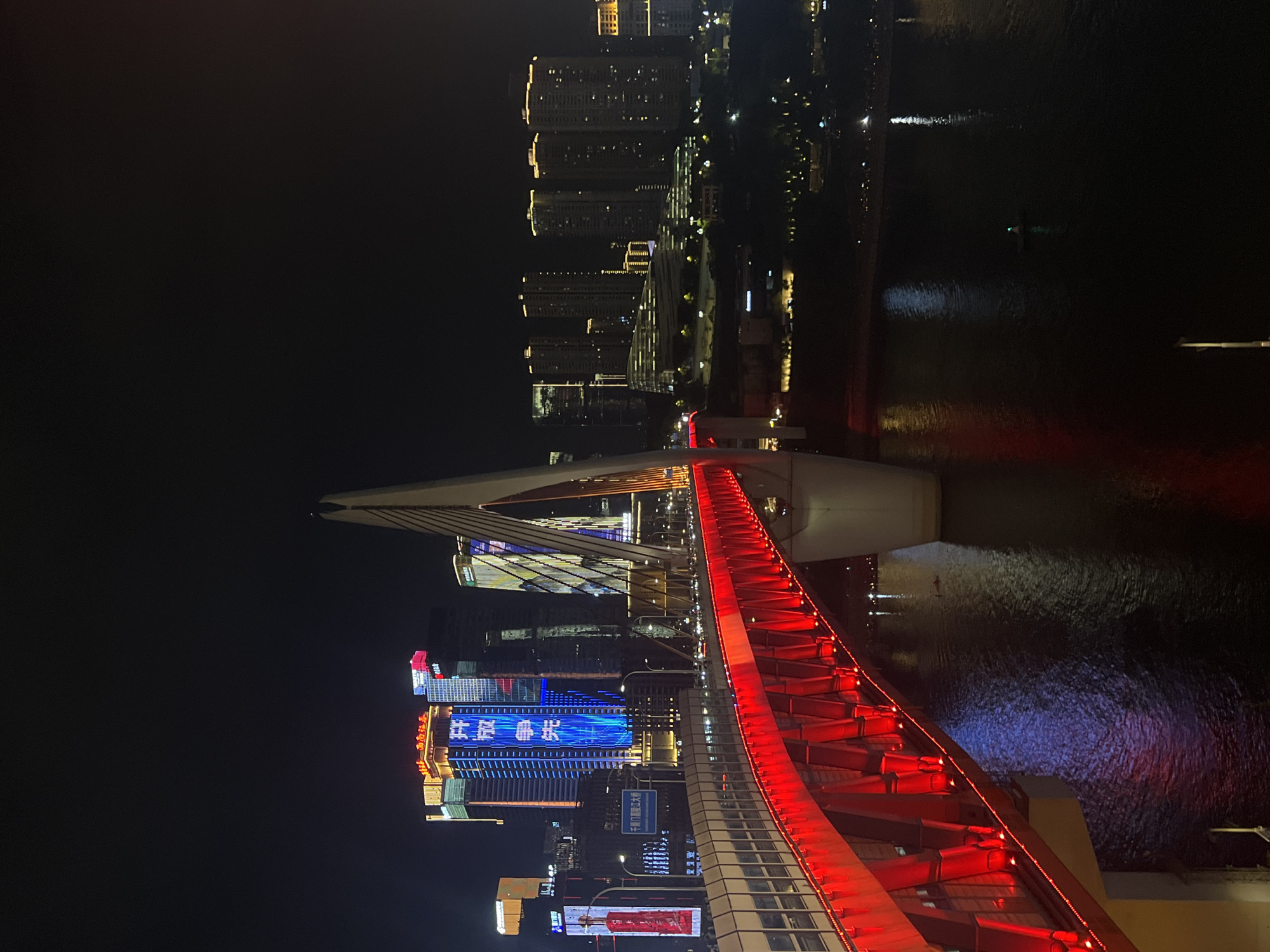 The image depicts the Hongyadong Bridge in Chongqing's Yuzhong District at night. The bridge is illuminated with vibrant red lights, creating a striking contrast against the dark sky and the water below. The cityscape of Yuzhong District is visible in the background, with numerous illuminated skyscrapers and buildings forming a glittering skyline. The bridge is a prominent structure, featuring a distinctive white pylon that extends upwards. There are vehicles visible on the bridge, suggesting it is a functional transportation artery. The water beneath the bridge reflects the city lights and the bridge's illumination, adding to the scenic night view. A small boat with a green light is visible on the dark water.