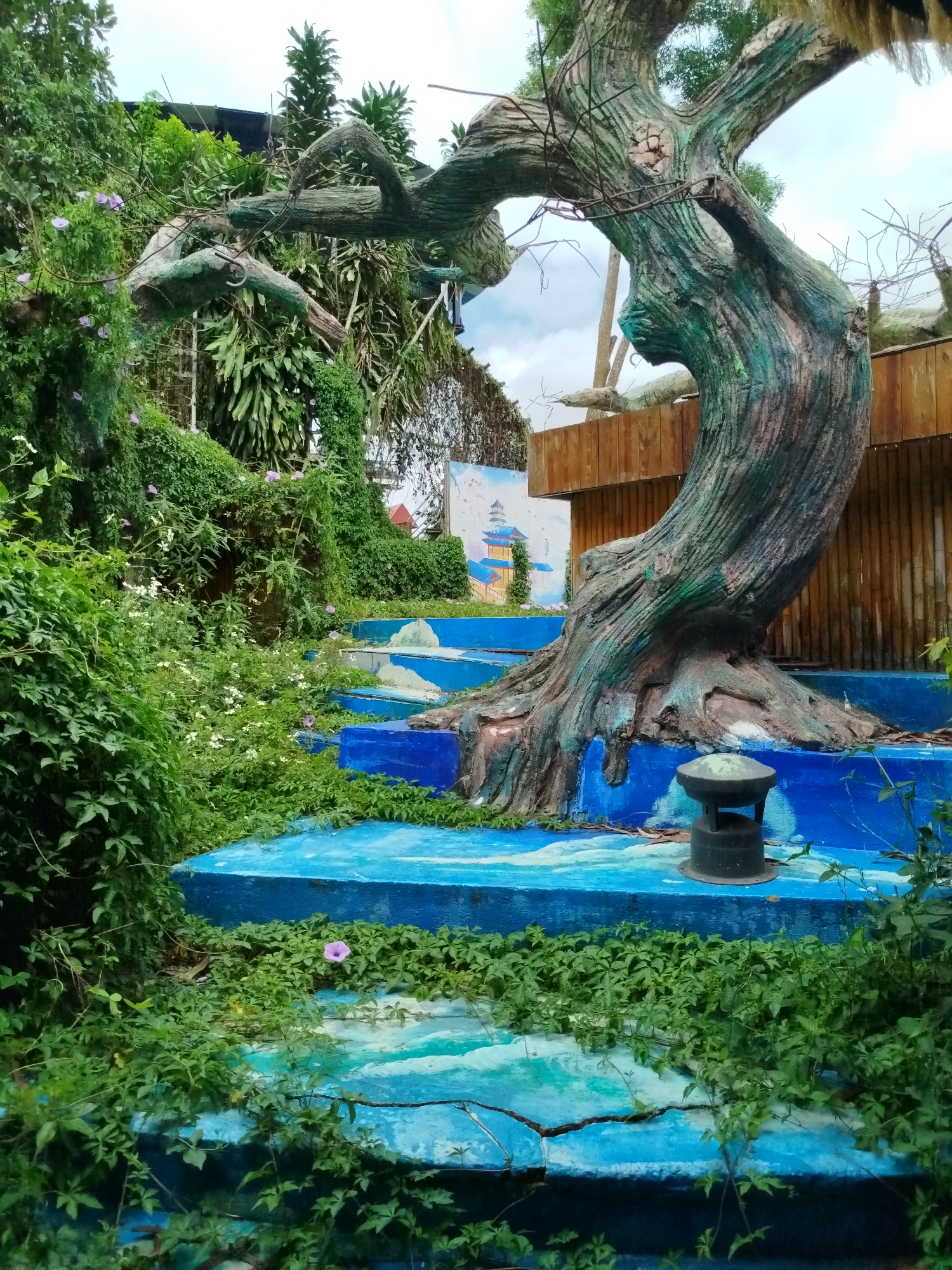 The image depicts an artificial tree sculpture, likely part of an outdoor attraction or garden in Da Lat, Vietnam. The sculpture is designed to resemble an aged, gnarled tree with textured bark painted in shades of green and brown. It stands on a multi-tiered platform, painted a vibrant blue and decorated with abstract white patterns that suggest water or clouds.
The platform is partially overgrown with lush green foliage and small white and purple flowers, indicating a humid, tropical environment. The sculpture itself has multiple branches, some of which are bare and spindly, while others are thick and robust. A small, dark, lantern-like object is placed on one of the blue platforms.
In the background, a wooden fence made of vertical planks is visible. Behind the fence, a mural painted on a wall depicts a traditional East Asian pagoda with blue roofs and yellow accents, set against a pale sky. The overall scene suggests a whimsical, perhaps slightly neglected, garden setting. No people are visible in the image. The lighting is soft, indicating an overcast or cloudy day. The ground beneath the blue platforms appears to be covered in fallen leaves and debris.