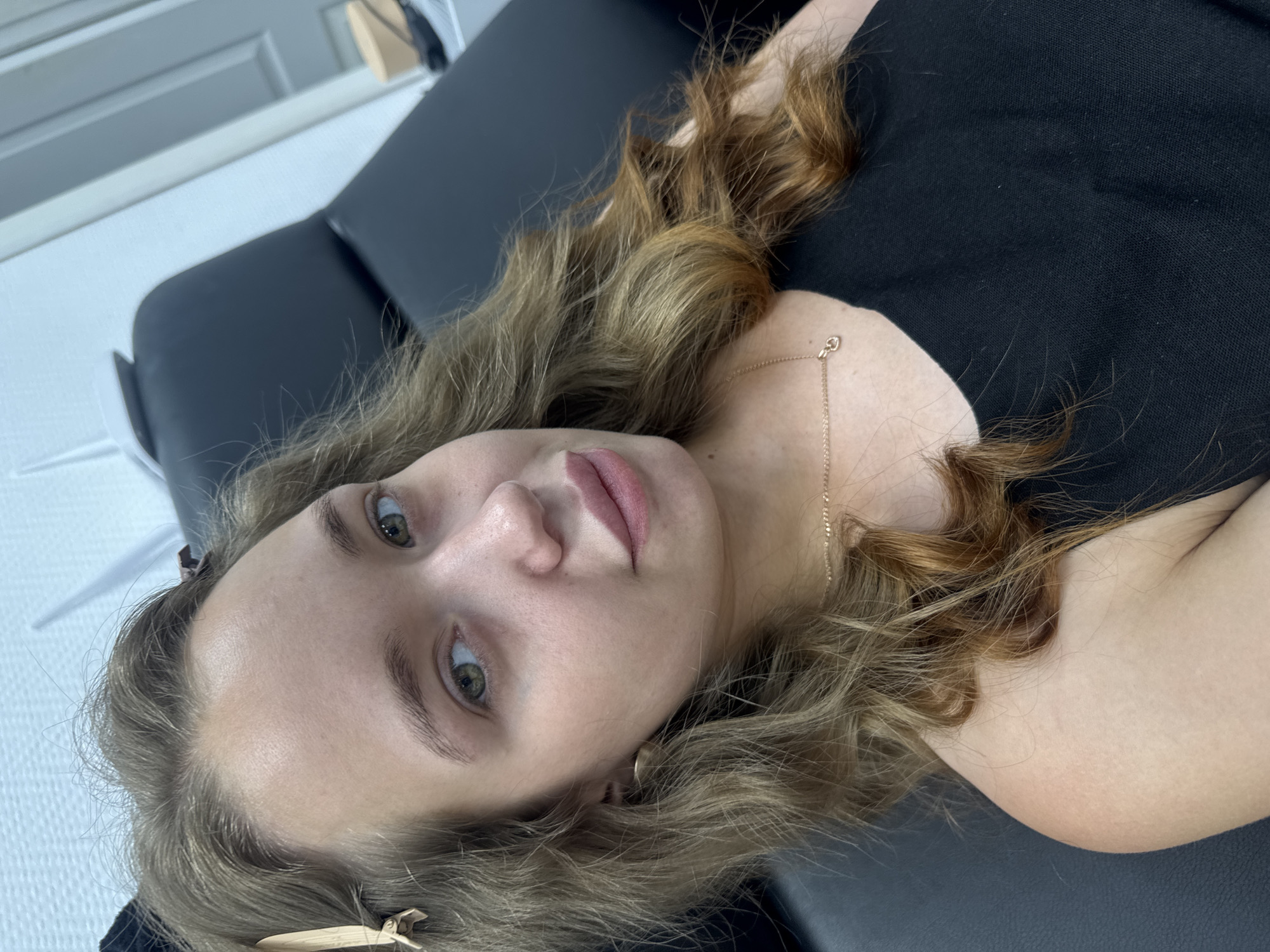 A young woman with light brown, wavy hair is looking directly at the camera. She is wearing a black top and a delicate gold necklace. Her hair is partially pulled back with a light brown clip. She appears to be indoors, possibly in a salon or waiting room, as indicated by the black chair she is sitting in and the light-colored wall behind her. The overall lighting suggests it is daytime. The image is a close-up portrait, focusing on her face and upper body. There is no identifiable information about her location within the city of Chaikovsky, Russia.