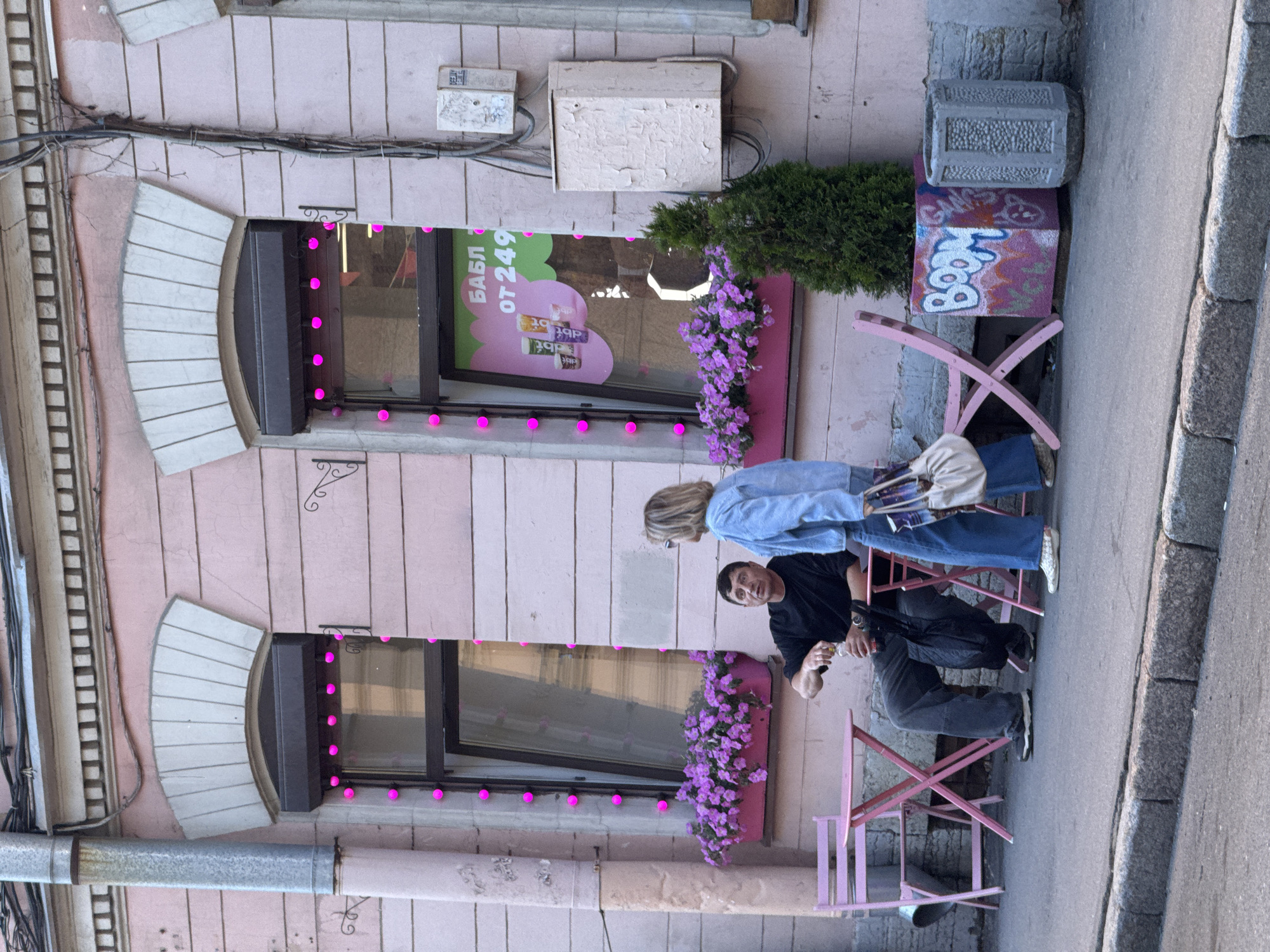 This image captures a street scene in Saint Petersburg, Russia, with two people interacting outside a building. The building features a pink facade with two large windows adorned with pink string lights. The upper window displays text that appears to be in Cyrillic, reading "БАБЛ ОТ 249", suggesting a promotion or pricing information. Below the windows, planters filled with vibrant purple flowers add a touch of color to the setting.
A woman with blonde hair, wearing a light blue denim jacket and blue jeans, stands facing away from the viewer. She carries a light-colored purse over her shoulder. Beside her, a man with dark hair, dressed in a black t-shirt and dark pants, is seated, looking towards the woman and holding a bottle. They are positioned near pink outdoor chairs and a small table, indicating a possible cafe or seating area.
In the background, a small evergreen bush and a planter box with graffiti on it are visible. The overall lighting suggests it is daytime, with overcast weather. The scene is set on a paved street with a cobblestone border, typical of urban environments. The architecture and the presence of Cyrillic text strongly indicate a location in Russia. The interaction between the two individuals appears casual and relaxed.