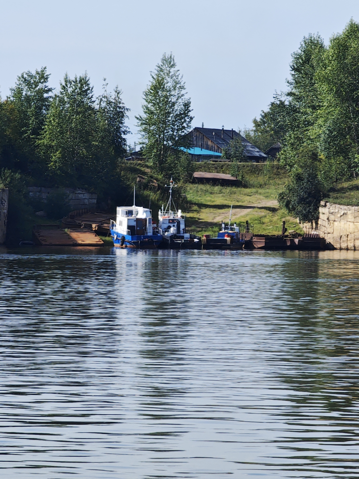 The image shows a body of water in front of a tree-lined bank. On the bank, there are several boats docked. In the background, there are some wooden buildings with dark roofs, partially obscured by trees. The sky is clear and blue. The location is the Vop River in the city of Yartsevo, Russia. The Vop River is a tributary of the Desna River and flows through the Smolensk Oblast. Yartsevo is a city in Smolensk Oblast, Russia, located on the Vop River. The river is a major waterway in the region and is used for transportation and recreation.