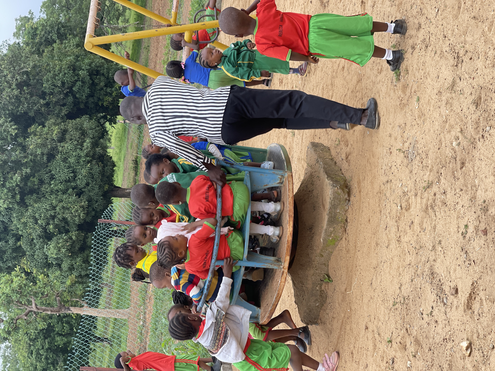 This image captures an outdoor playground scene in Akwanga, Nigeria, showing a group of young children and one adult male engaged in playtime activities. The ground is sandy dirt, and lush green trees and vegetation form the background.
In the foreground and midground, a blue and brown metal merry-go-round is visible, with several children seated on it. An adult male, wearing a black and white striped long-sleeved shirt and black trousers, is standing next to the merry-go-round, appearing to supervise or push it for the children. Many of the children are wearing school uniforms; some are in red tops with green shorts or skirts, while others wear green and yellow tracksuits or blue tops with green bottoms. Their hair is often styled in braids.
In the background, a yellow metal swing set can be seen, with other children standing near or on it. One child, wearing a red top and green shorts near the swing set, has the text "REBECA HOU" (likely "REBECCA HOUSE") visible on their shirt. A chain-link fence separates the play area from a more overgrown section of trees and greenery.
The scene depicts a daytime setting with natural light, suggesting pleasant weather for outdoor play. The overall atmosphere is one of active supervision and children enjoying recreational time, likely during a school break or a community event.