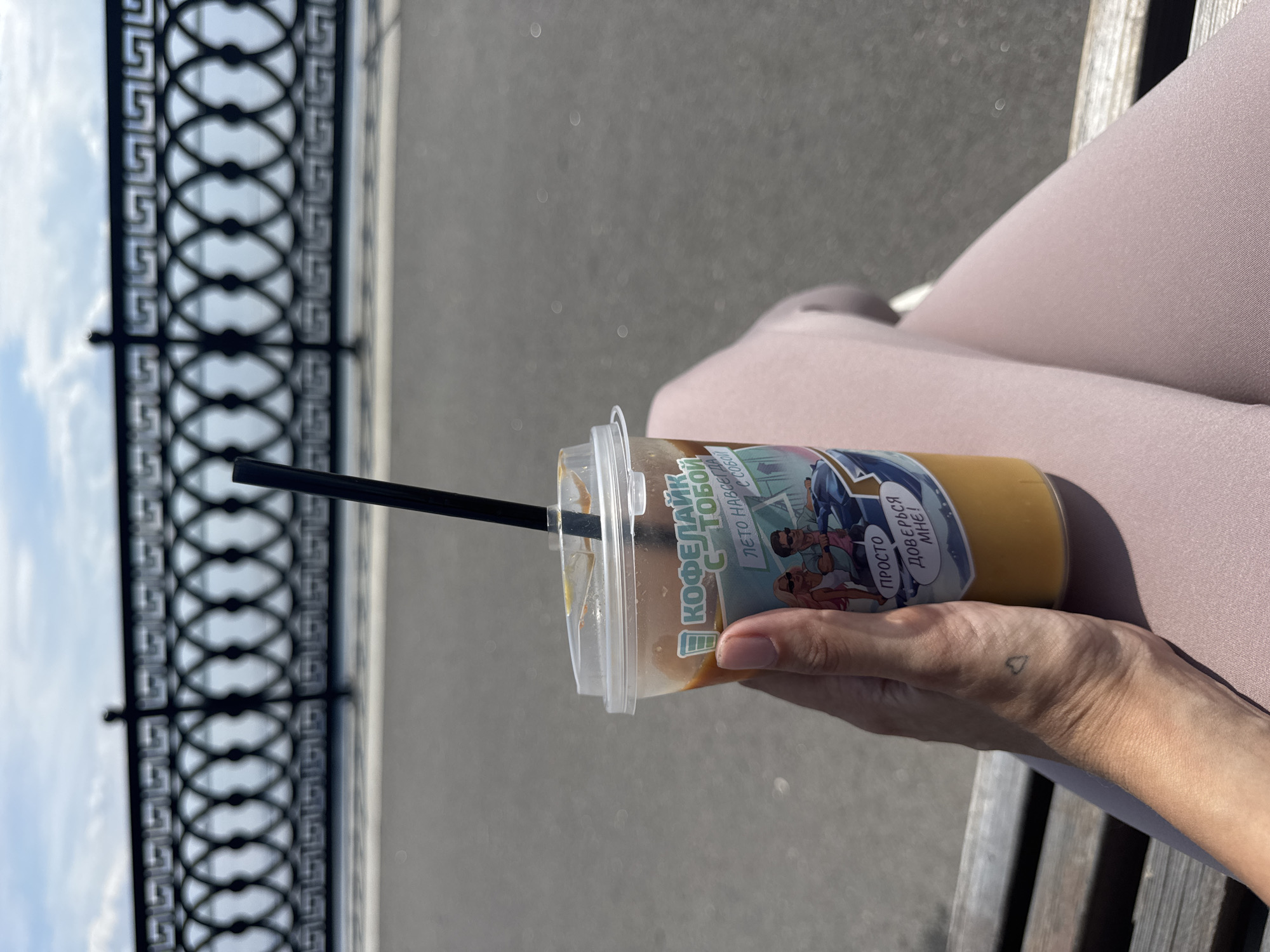 The image captures a close-up view of a person's hand holding a clear plastic cup filled with a light brown beverage, likely coffee or tea, and a black straw. The cup has a colorful sticker with comic-style illustrations. Visible text on the sticker includes "КОФЕЛАЙК" and "ЛЕТО НАВЕ", suggesting a summer theme related to a coffee brand. Another phrase, "ПРОСТО ДОВЕРЬСЯ МНЕ!", translates to "JUST TRUST ME!". A small heart tattoo is visible on the wrist of the hand holding the cup.
The background suggests an outdoor setting with a blurry grey surface, possibly pavement or a pathway. To the left, a black decorative metal railing with an intricate geometric pattern is partially visible against a bright, overcast sky. The overall lighting indicates daytime, and the presence of the railing and the sky suggests the person is likely seated on a bench outdoors, possibly near a promenade or park, given the context of Saratov, Russia. The image conveys a relaxed and casual atmosphere, perhaps a moment of enjoying a drink on a warm day.