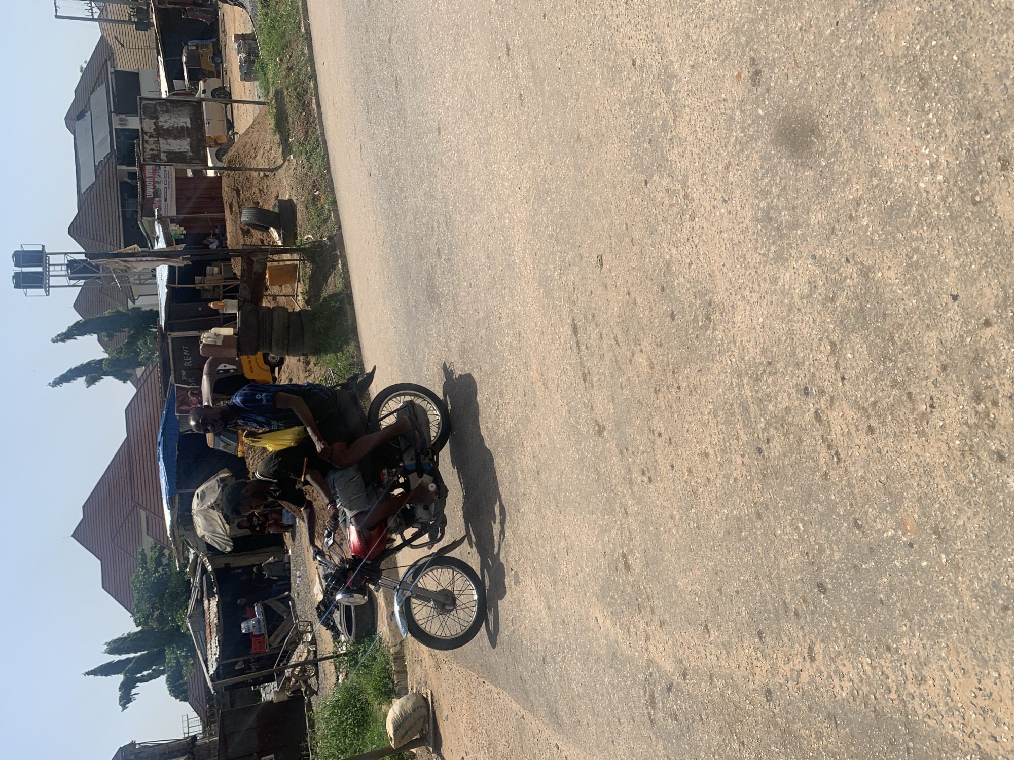 The image depicts a street scene in Abuja, Nigeria. A motorcycle with two male riders is moving on a paved road. The rider in front is wearing a blue shirt, while the passenger in the back is wearing a yellow shirt. The roadside features informal structures, including makeshift shops or stalls. In the background are buildings with corrugated metal roofs. Also visible is a water tank. The road surface is worn. Vegetation is sparse.