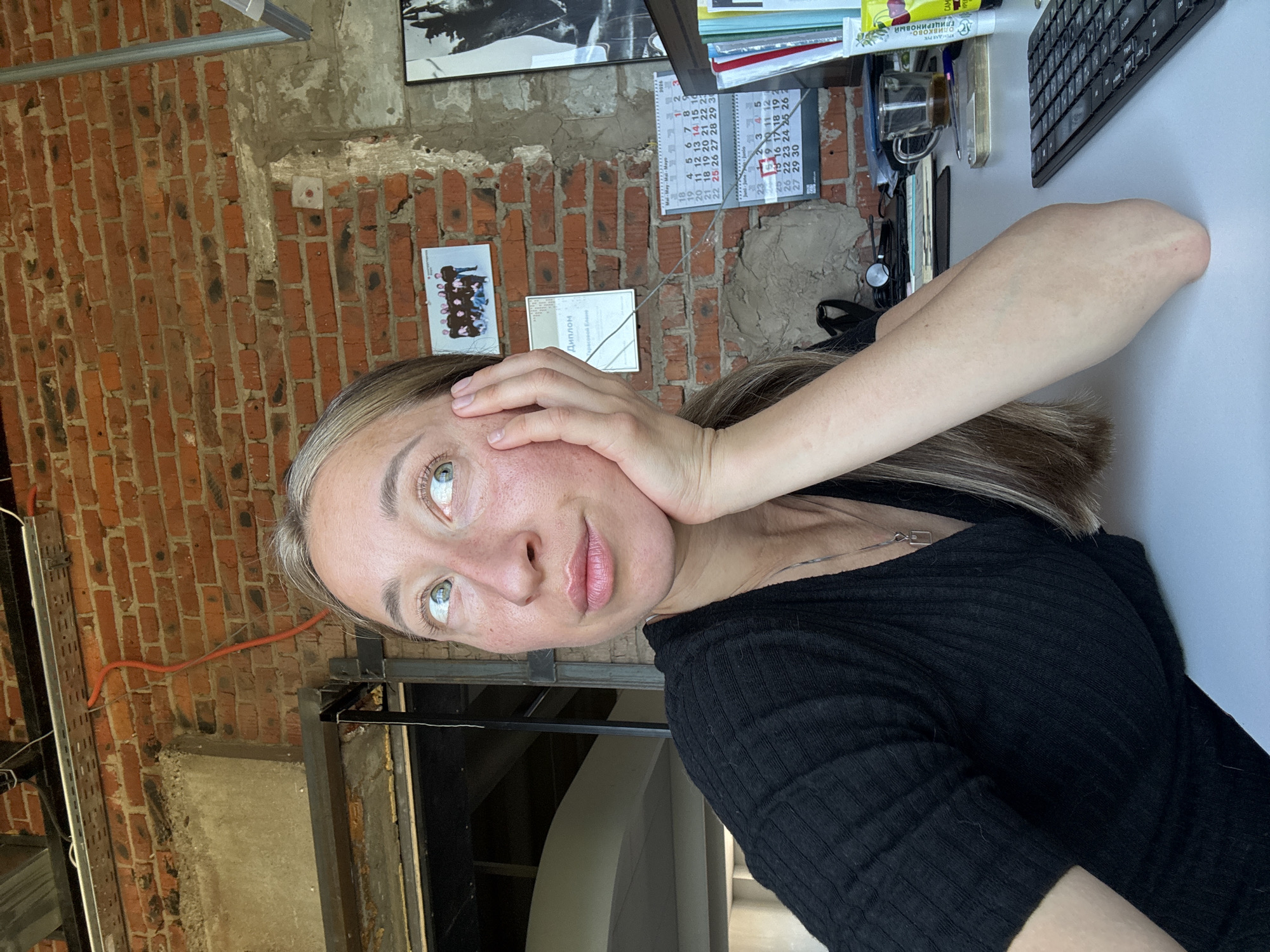 A woman with blonde hair, wearing a black ribbed top and a silver necklace, rests her head on her hand as she looks up and to the right, appearing thoughtful. She is seated at a white desk in front of a brick wall. On the wall, there are two framed pictures, one a group photo and the other a certificate. Calendars are also visible on the wall. The desk has a keyboard, a mug of tea, and various office supplies. The environment appears to be an office or workspace with industrial elements like exposed brick and metal shelving. The location is identified as Chaikovsky, Russia.